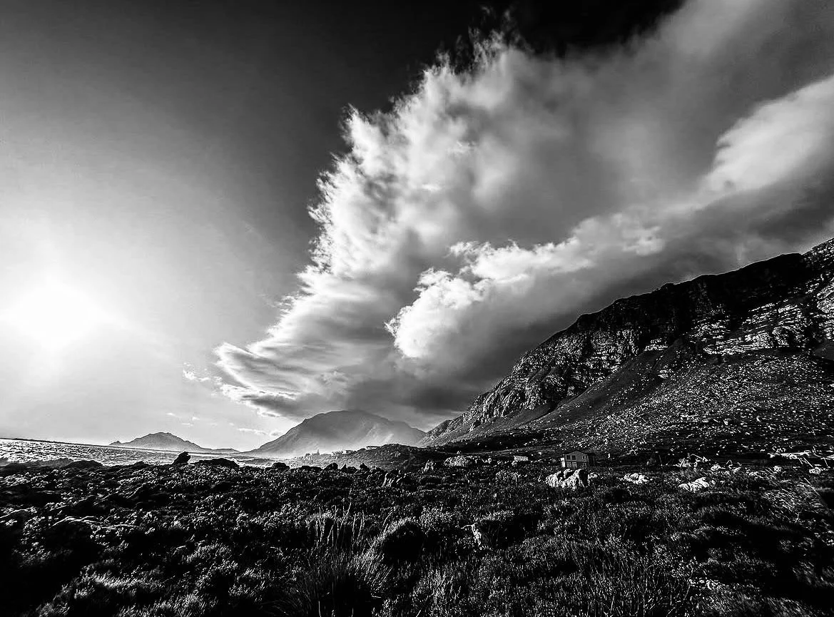 Black and white photograph of a rugged coastline with mountains in the background and dramatic clouds overhead.