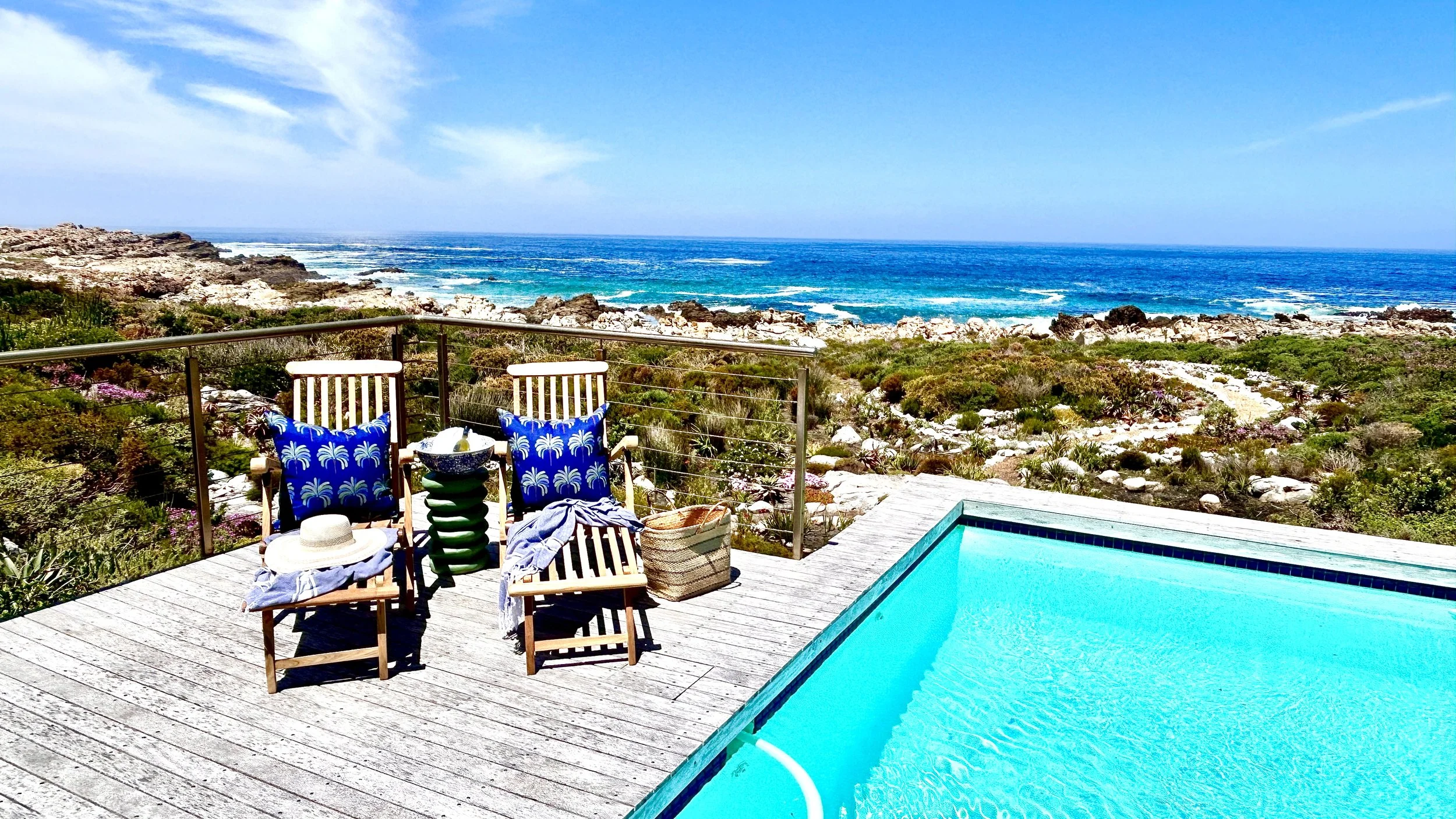 Deck with two wooden chairs and blue pillows with palm tree pattern, a straw hat, and a striped bag, overlooking a rocky coastline and the ocean, with a swimming pool in the foreground.