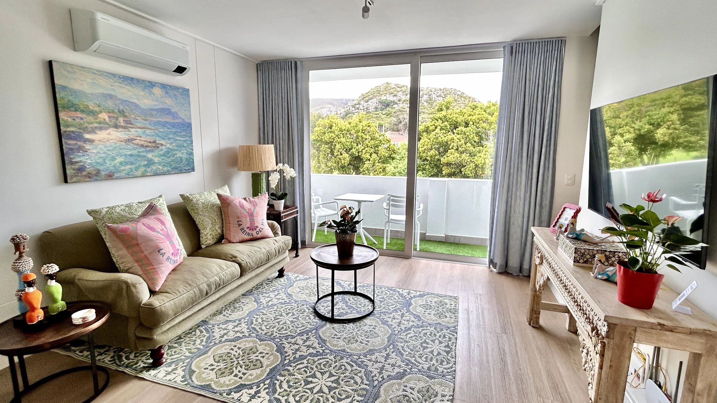 Living room with a beige sofa, pink pillows, a blue patterned rug, a wooden coffee table with potted plant, a large wall-mounted TV, a cream-colored carved console table with a potted plant, a large window with blue curtains, and a view of a balcony 