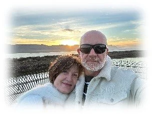 A man and woman taking a selfie outdoors at sunset near a body of water and mountains in the background.