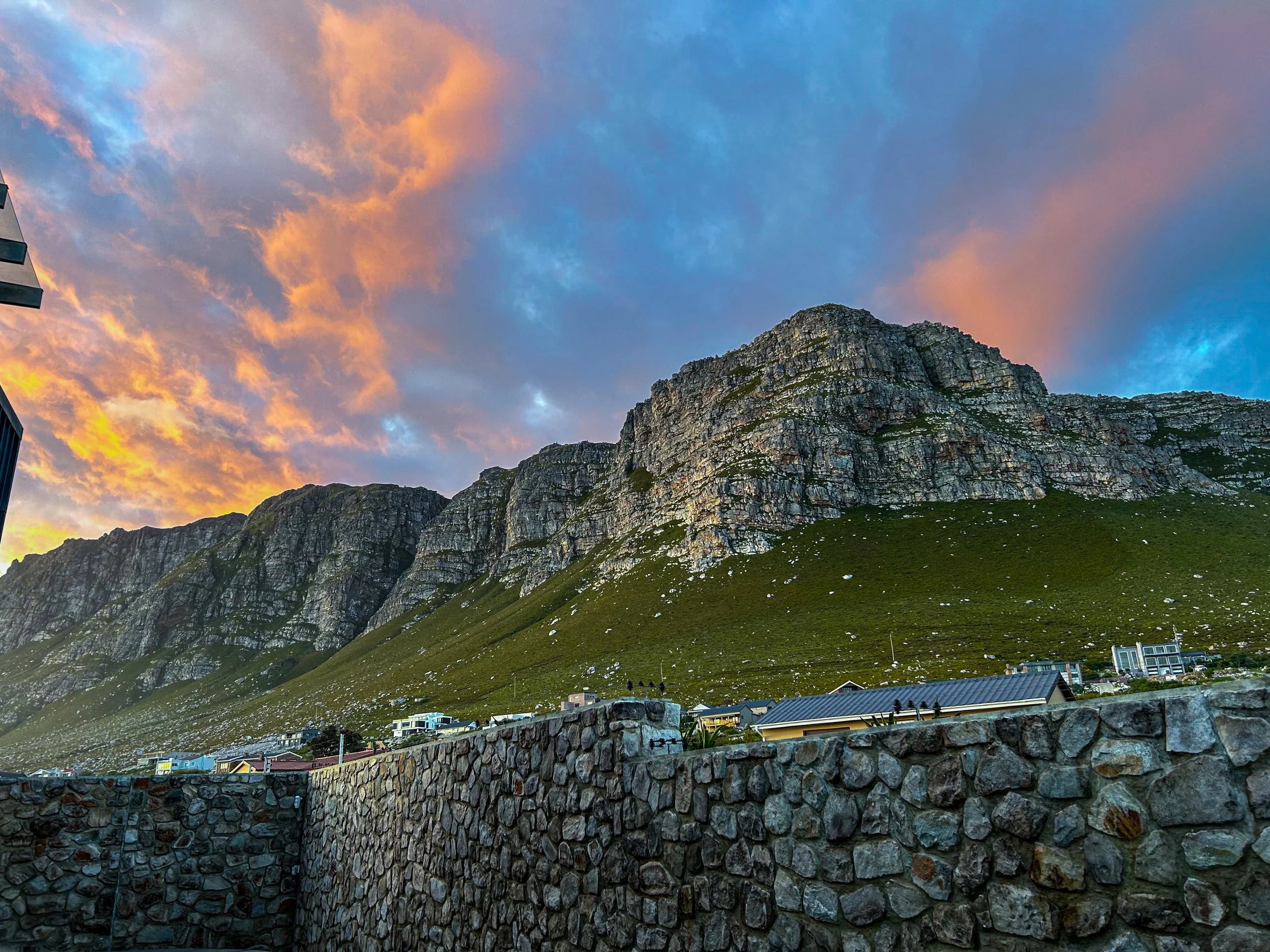 Mountain with green hillside, houses, and a stone wall in the foreground, under a colorful sunset sky.