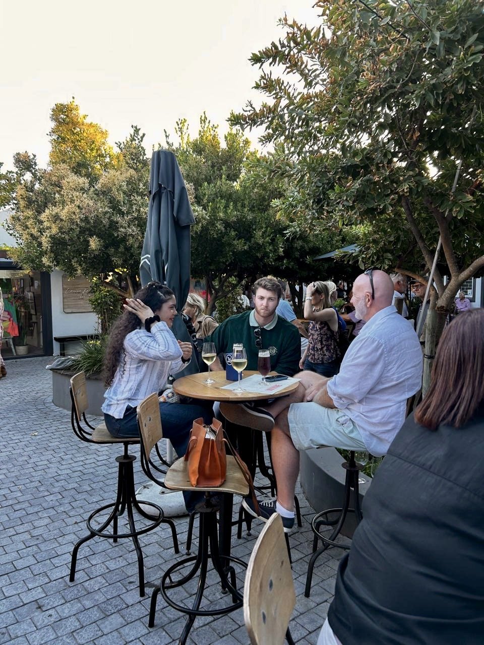 People enjoying drinks at an outdoor café, sitting around a small round table with drinks, surrounded by trees and a cobblestone patio.