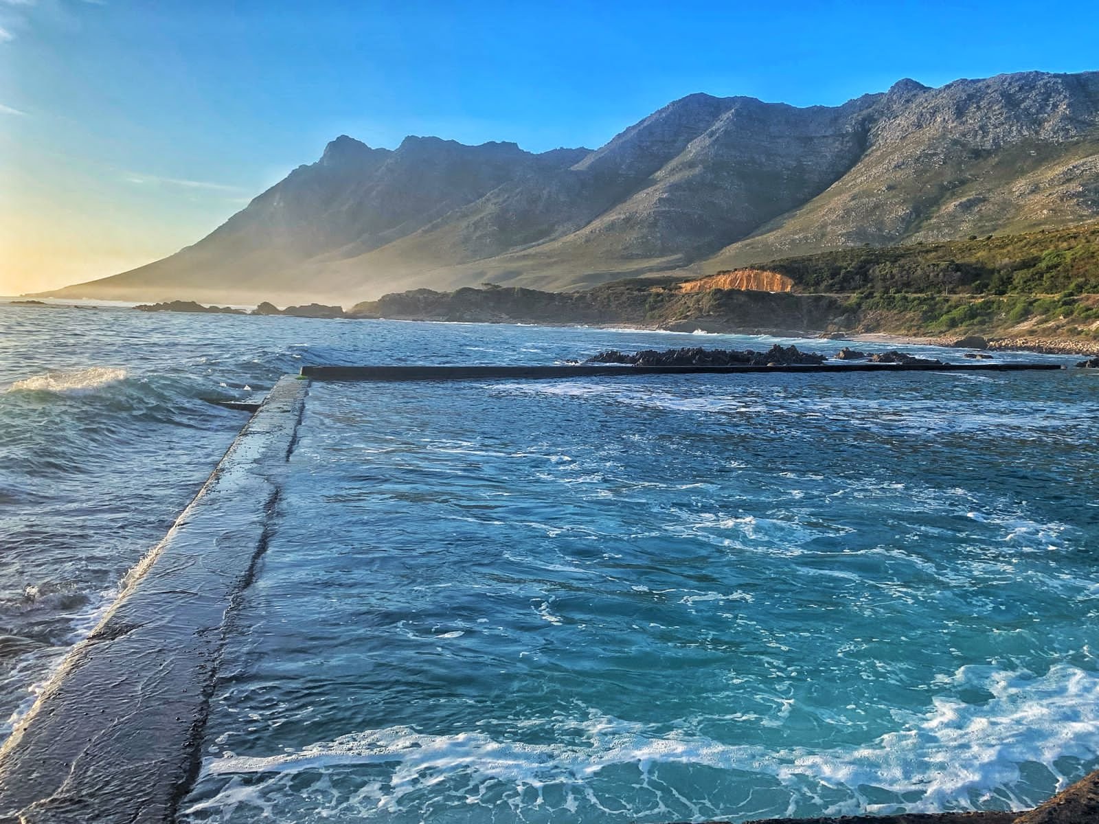 A scenic view of the ocean with mountains in the background, showing the edge of a swimming pool or natural pool along the rocky shoreline during sunrise or sunset.