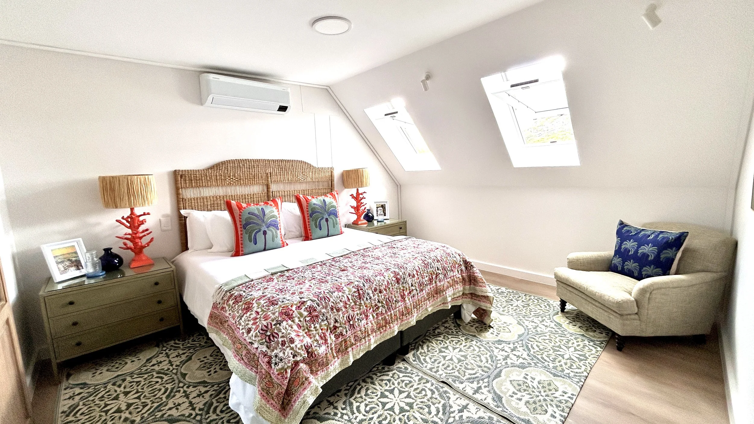 Bedroom with a large bed with colorful pillows, a woven headboard, side tables with lamps, a beige armchair with a blue pillow, and two skylight windows.