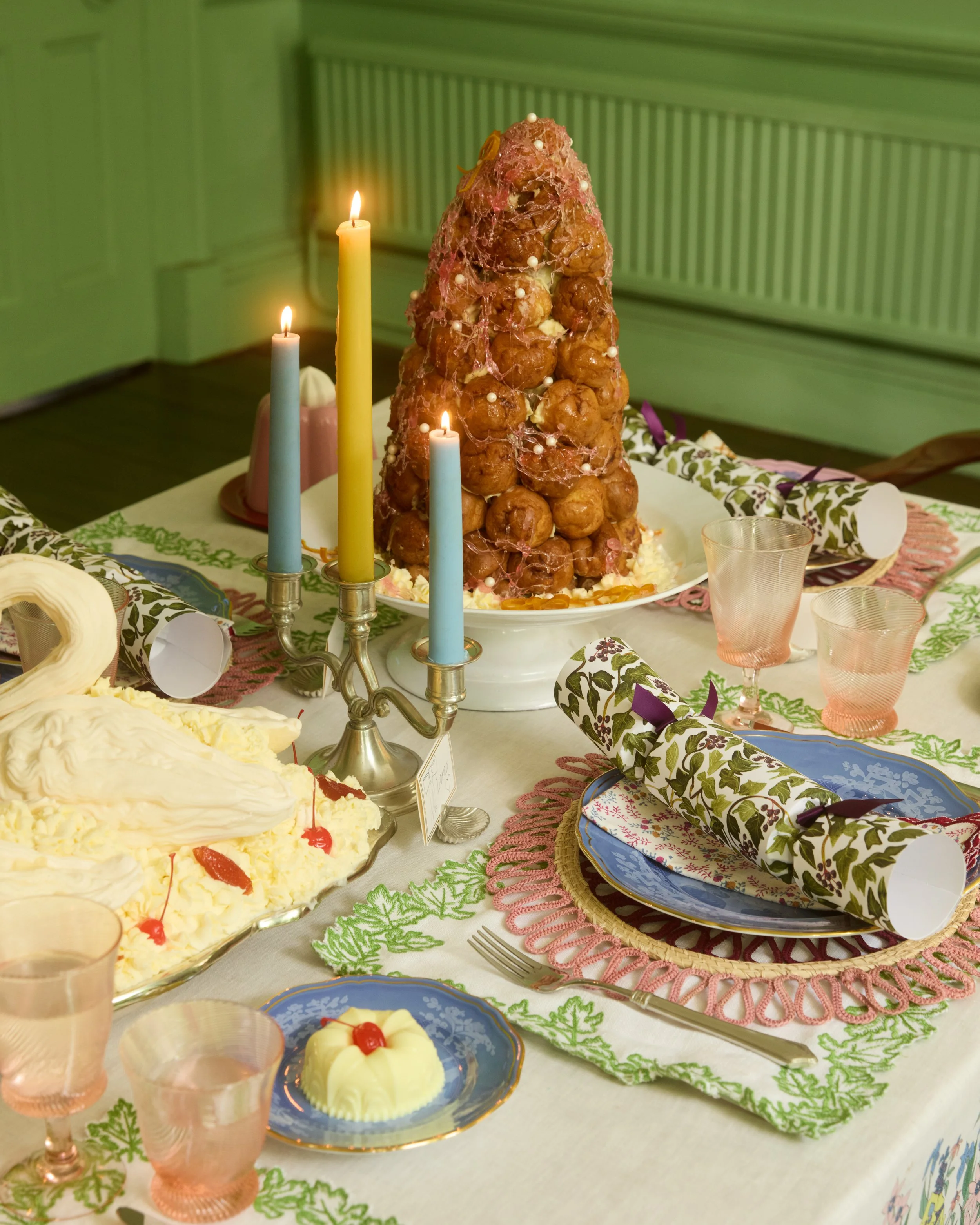 A Christmas or holiday dinner table with a croquembouche centerpiece, decorated with small white pearls and pink spun sugar, surrounded by various desserts, colorful candles, and patterned plates and napkins.