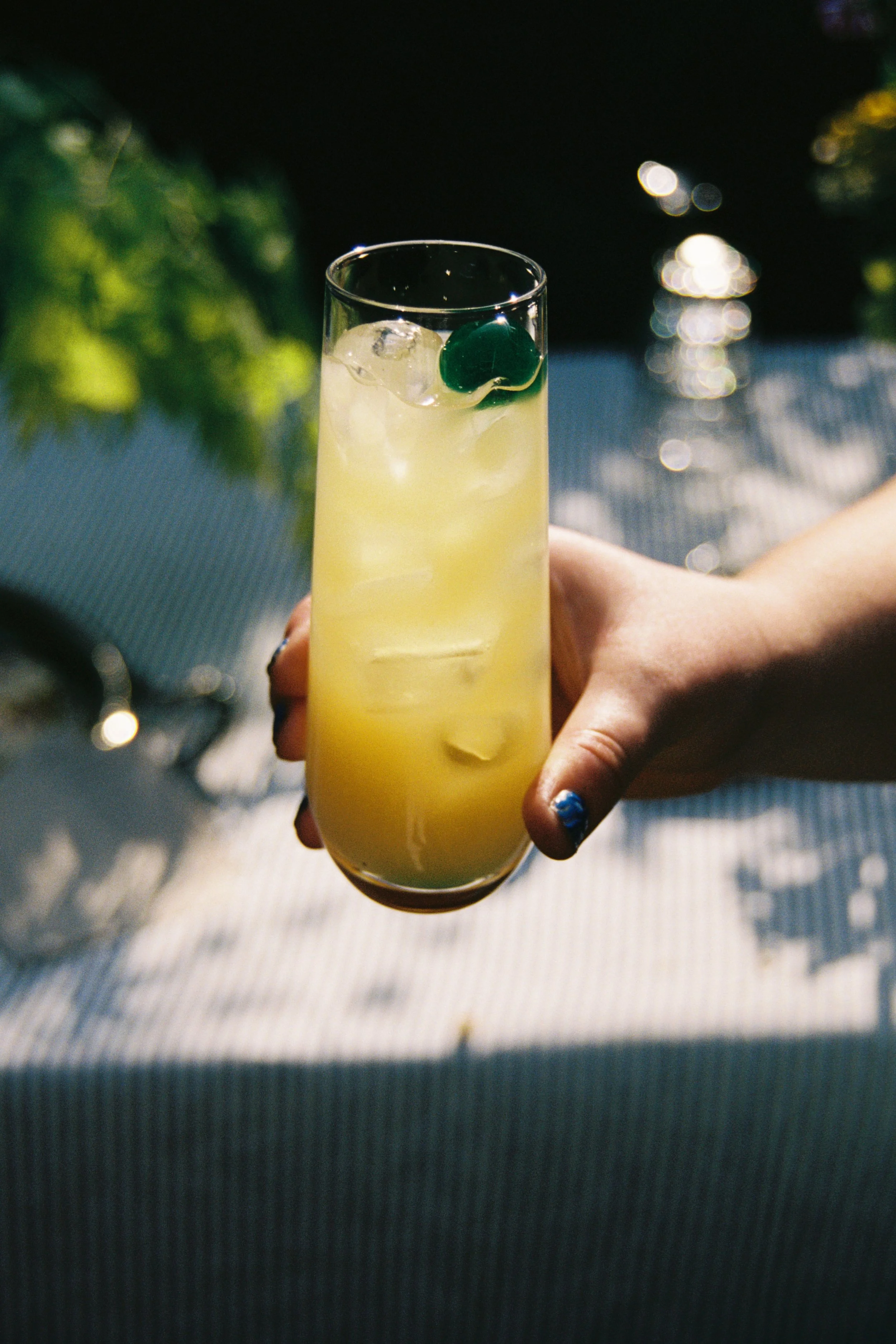 A hand holding a tall glass of yellow cocktail with ice and a small green fruit garnish, set outdoors on a sunny day.