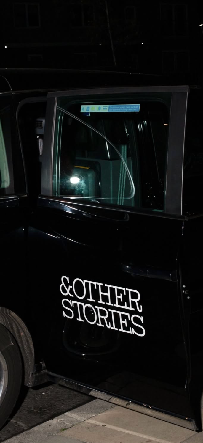 A black vehicle with the words '&OTHER STORIES' written on the door, parked beside another vehicle at night.