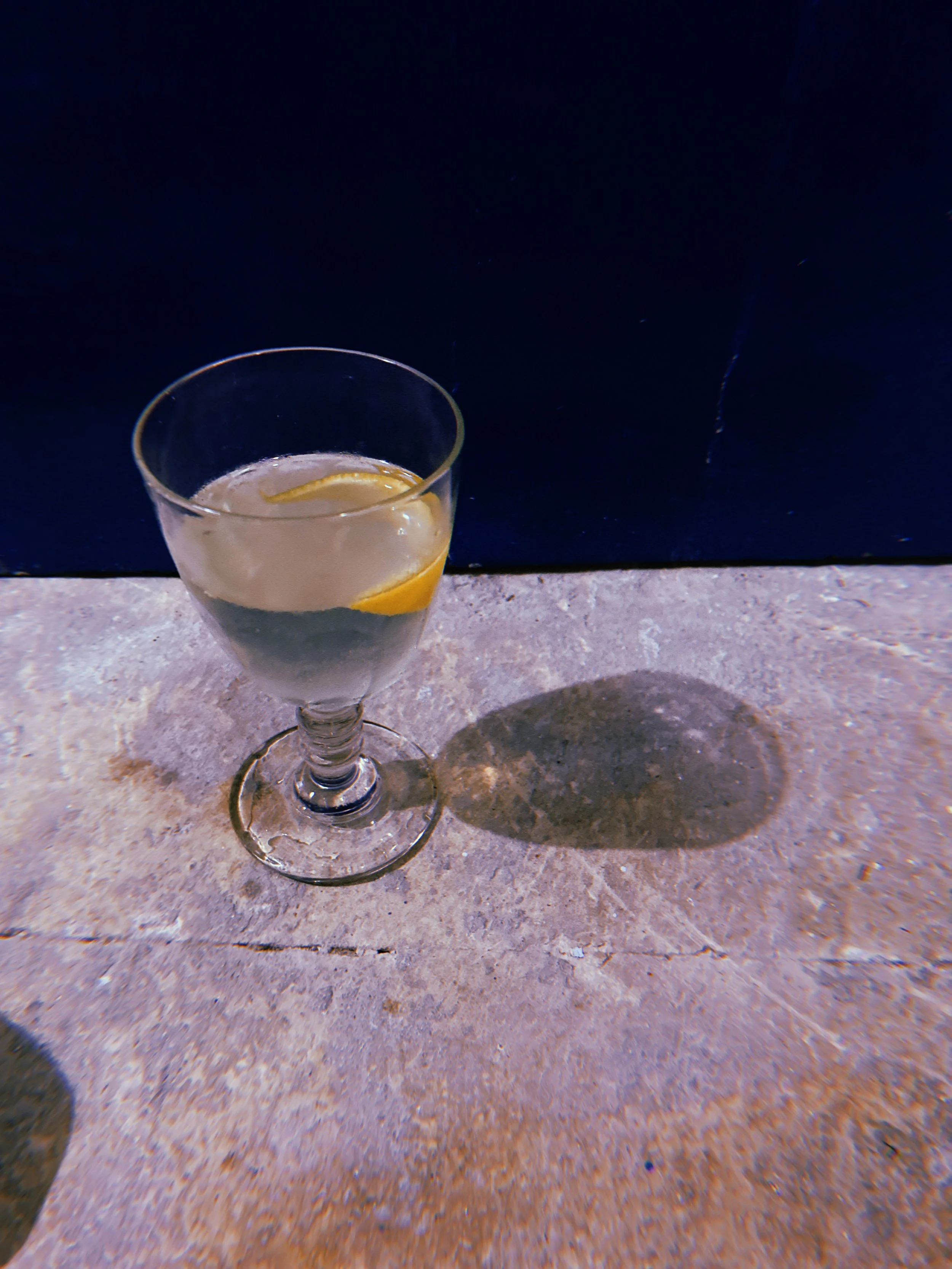A glass of water with lemon slices on a textured surface, casting a shadow on the ground.