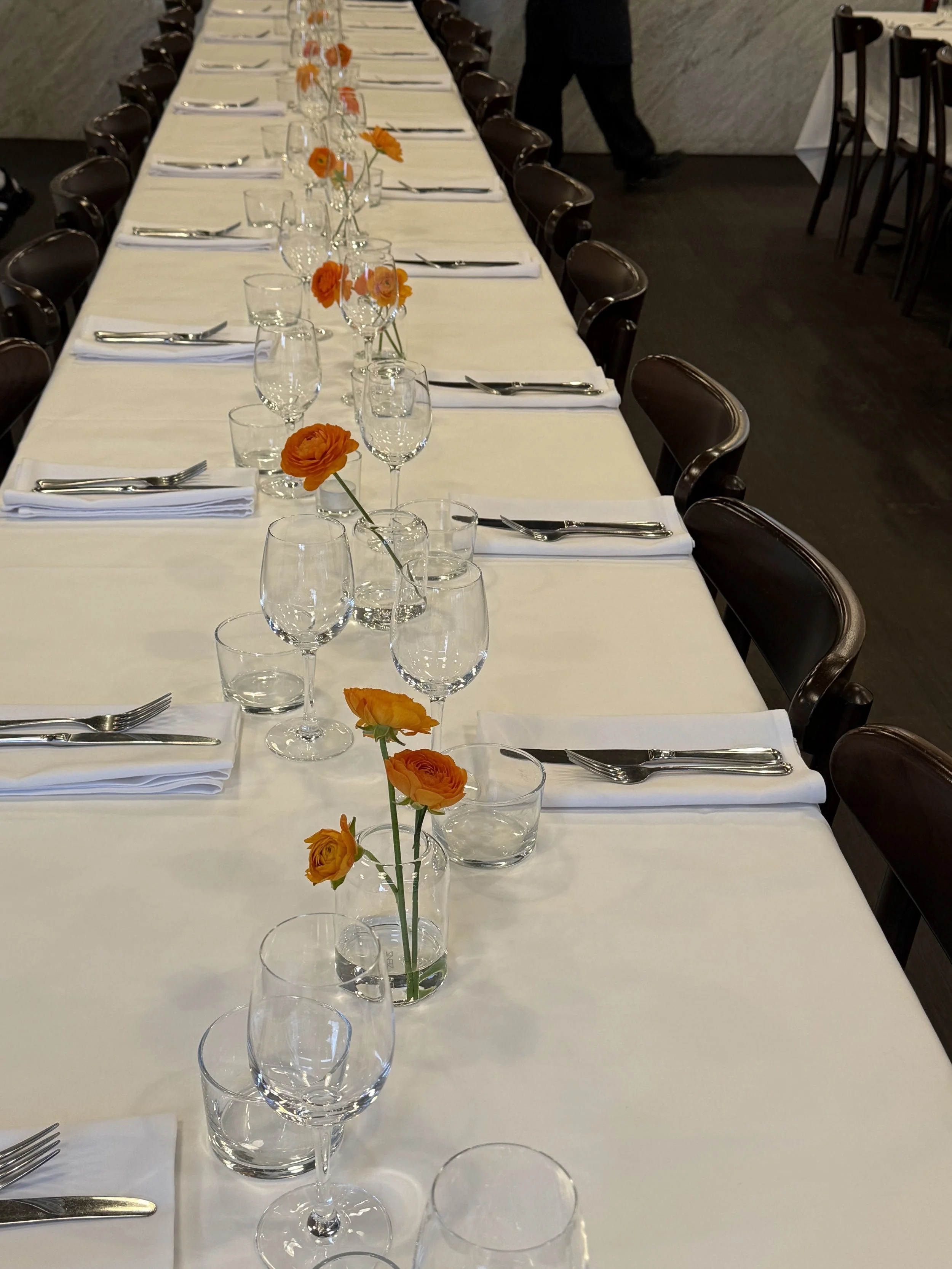 A long dining table set for a formal event with white tablecloths, orange flowers in small vases, wine glasses, water glasses, napkins, and cutlery.