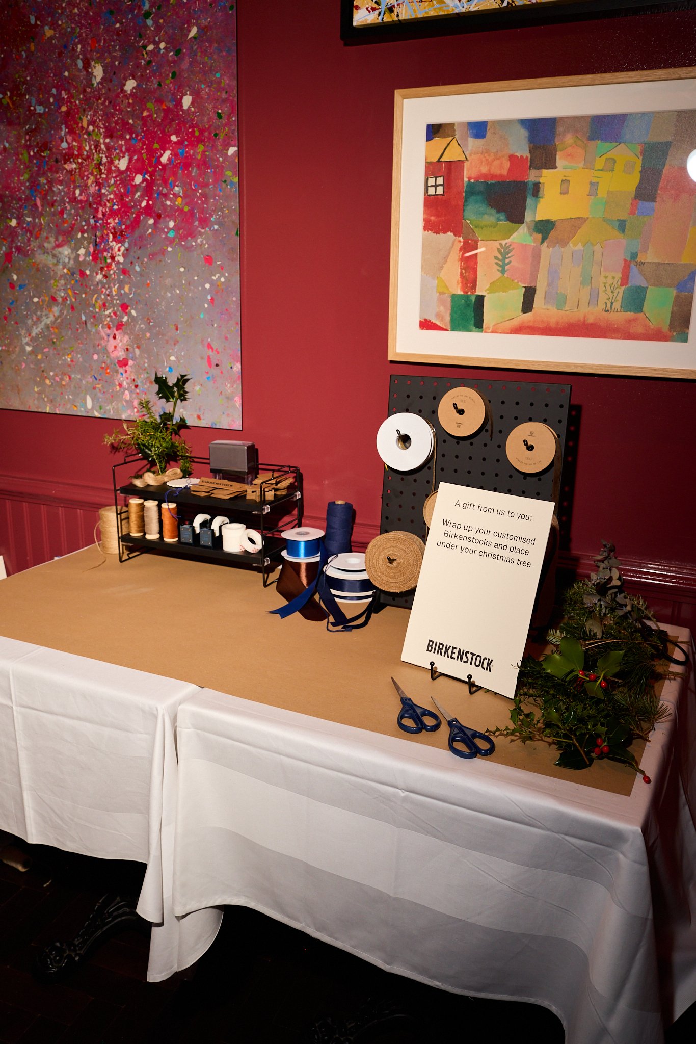 Table with green holiday foliage and red berries, scissors, a sign that reads 'A gift from us to you: Wrap up your customised Birkenstock and place under your Christmas tree,' and various craft supplies including thread spools and rolls of ribbon, ag