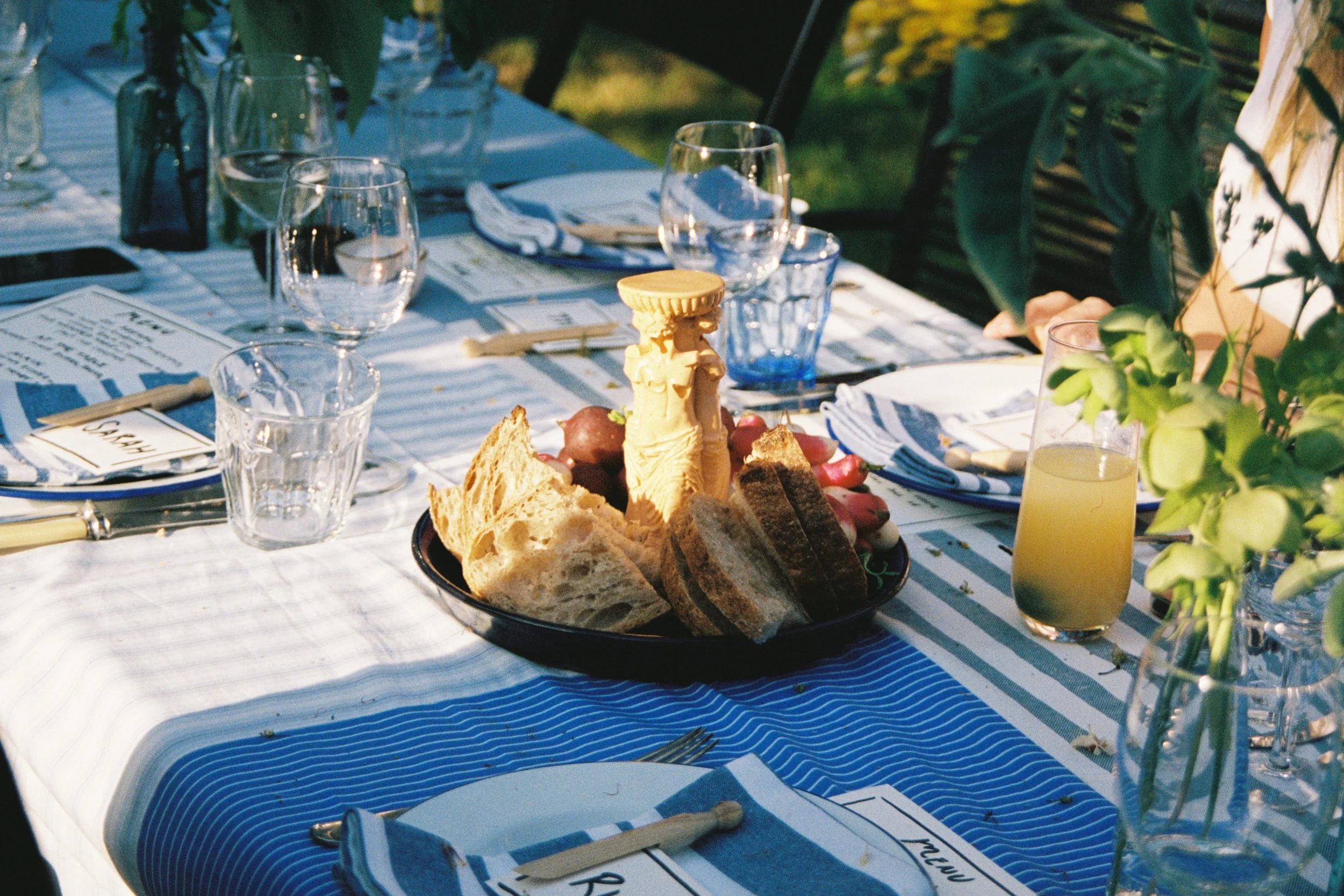 Table set outdoors with plates, glasses, napkins, and a black dish in the center containing bread, radishes, and a decorative statue. Greenery surrounds the scene.
