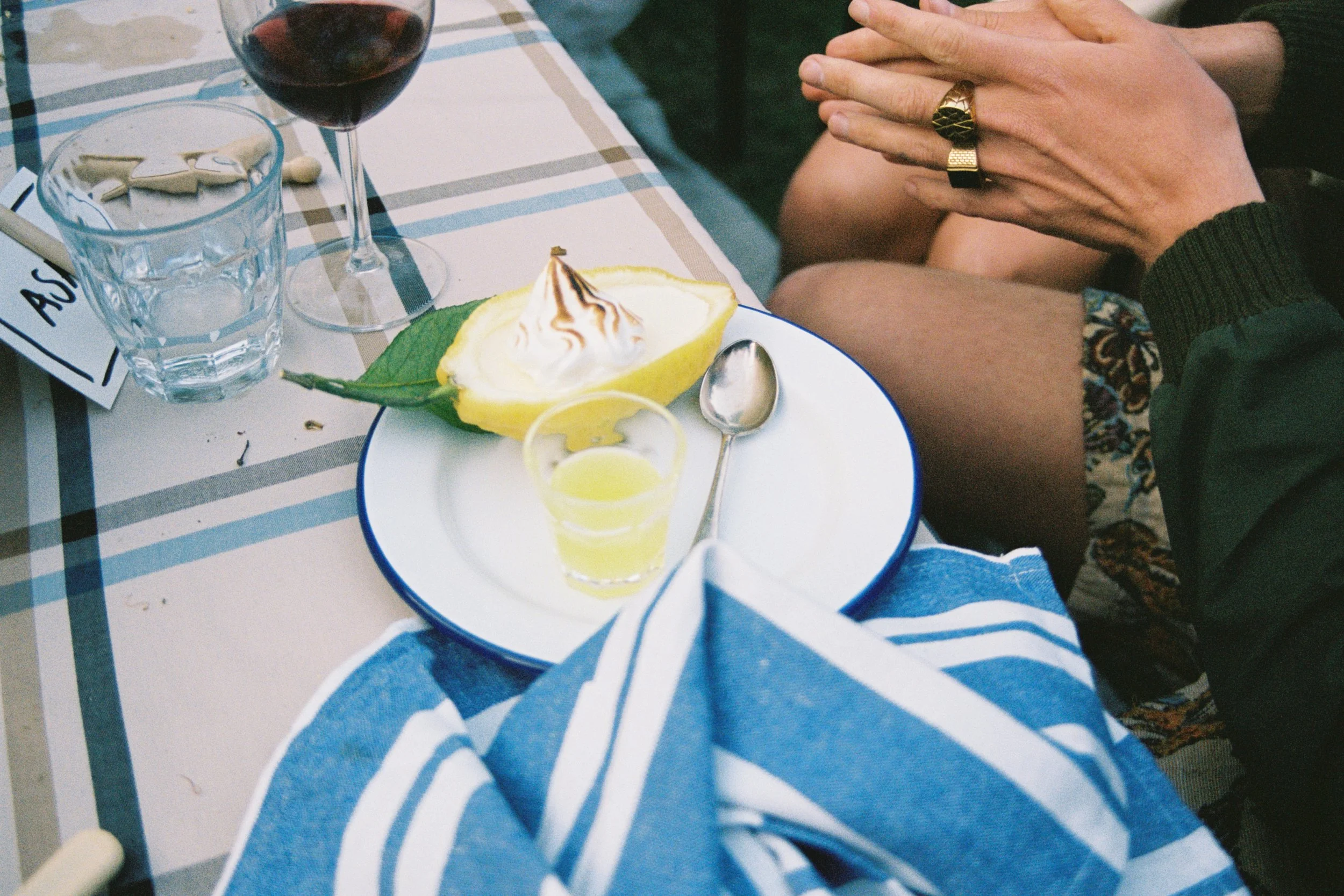 Partially eaten lemon with whipped cream and chocolate drizzle, small glass of lemon drink, spoon, wine glass with red wine, glass of water, tablecloth, person's hands with rings resting on knee, blue and white striped cloth, and part of a dining set