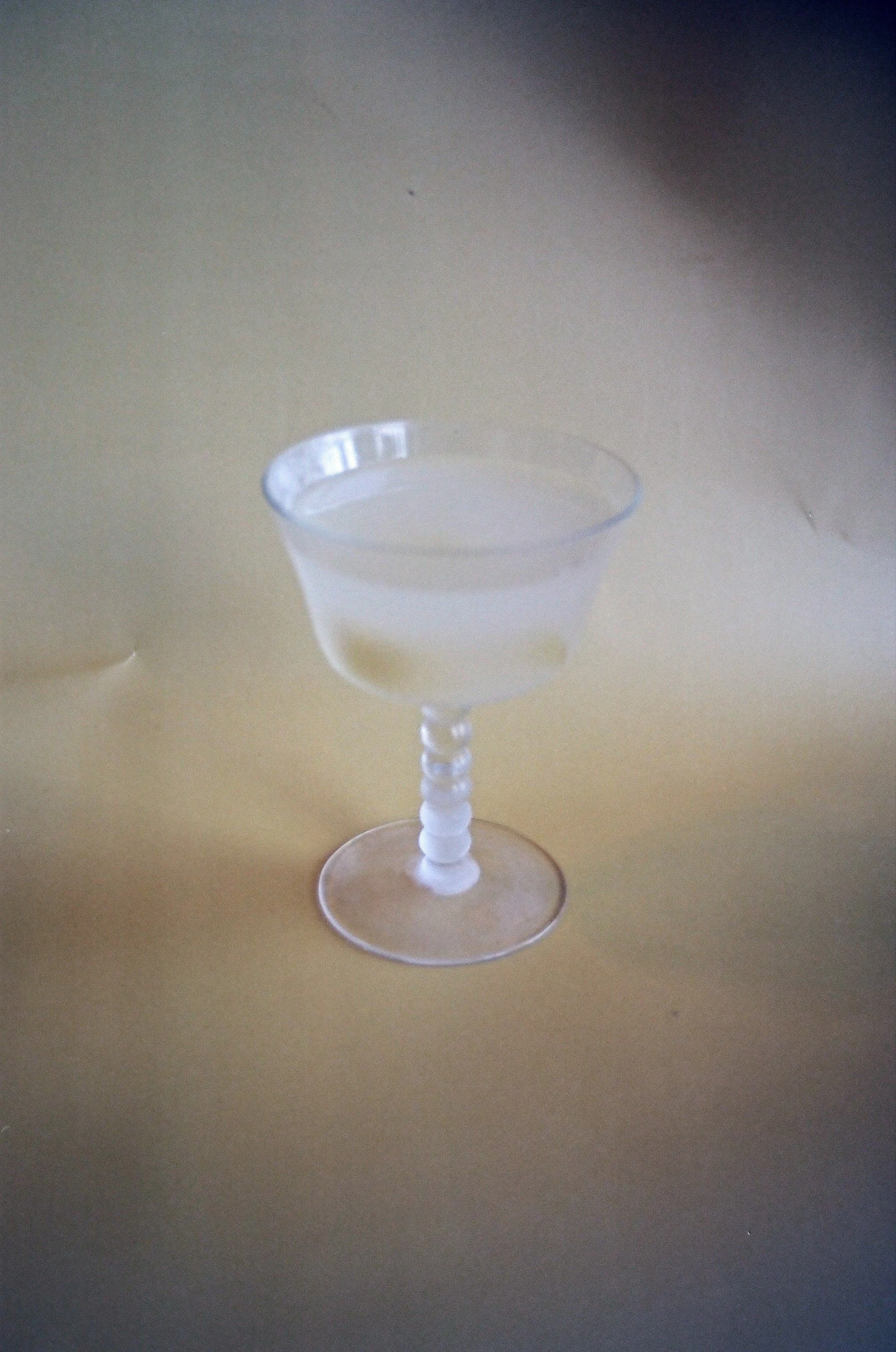 A clear cocktail glass with a decorated stem, filled with a transparent liquid, placed on a beige surface.