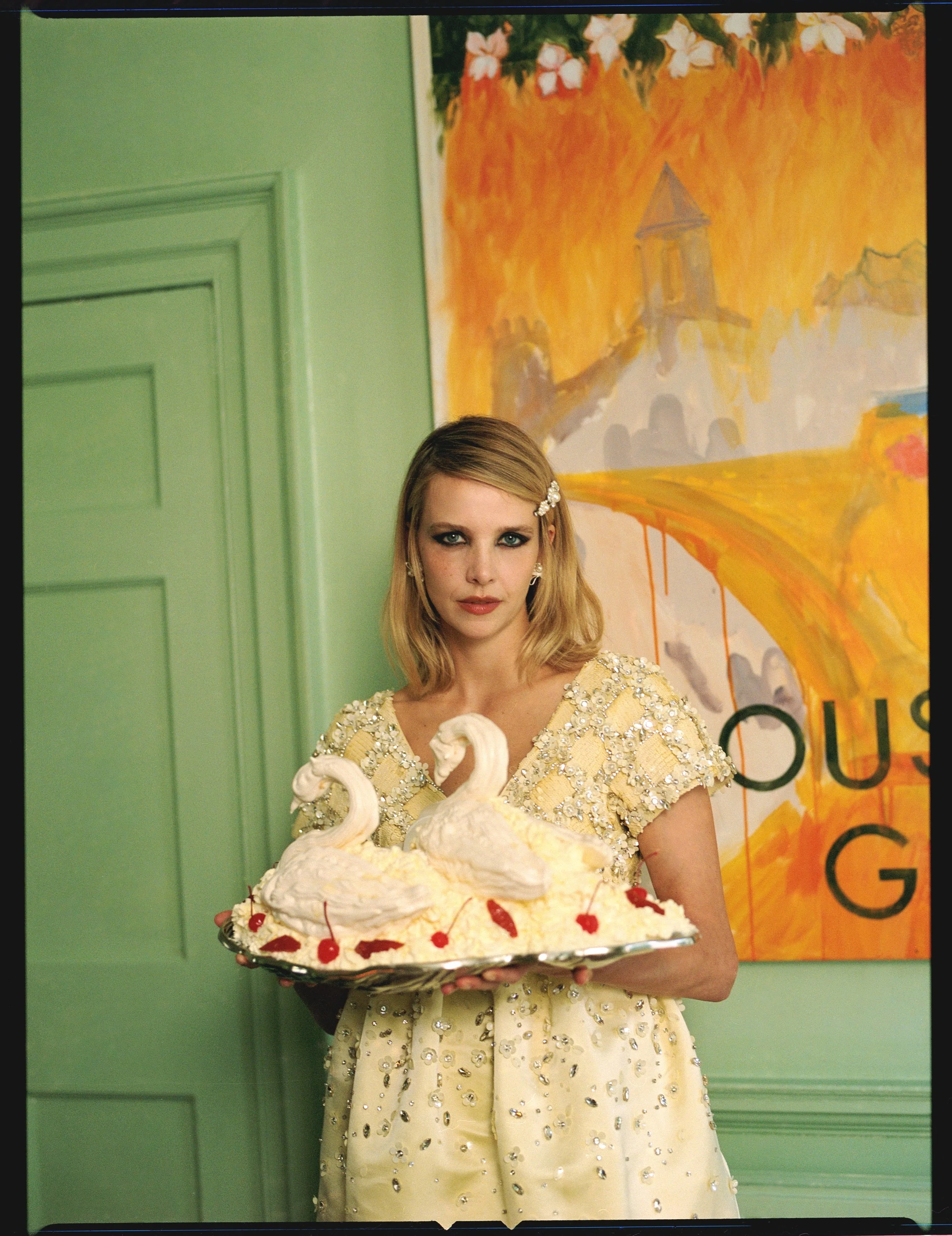 A woman in a yellow dress decorated with sequins, holding a plate with a cake shaped like swans, topped with cherries, in a room with green walls and a colorful painting in the background.
