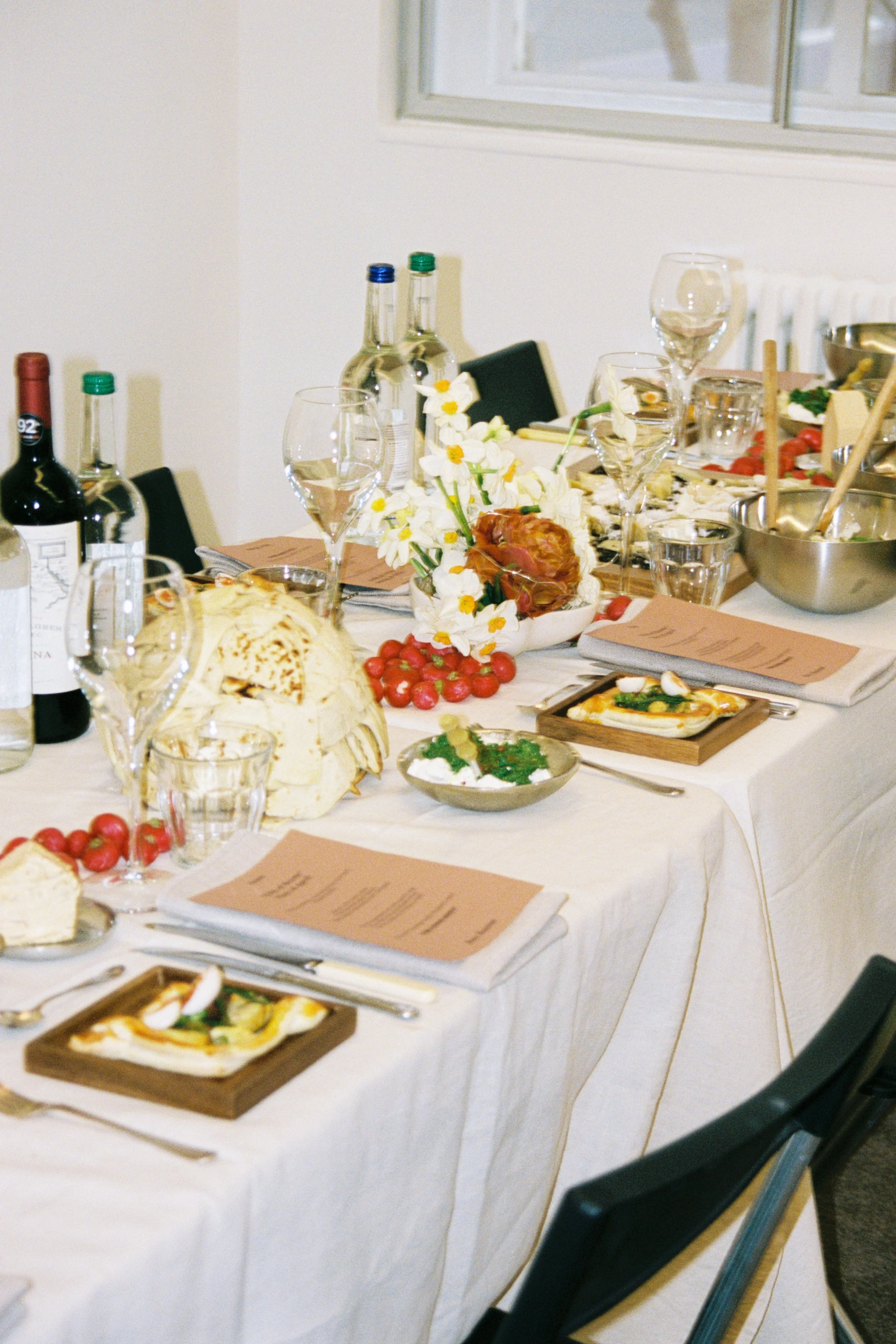 A decorated dining table set for a meal with wine bottles, glasses, floral centerpiece, bread, cherry tomatoes, salad, and menu cards.