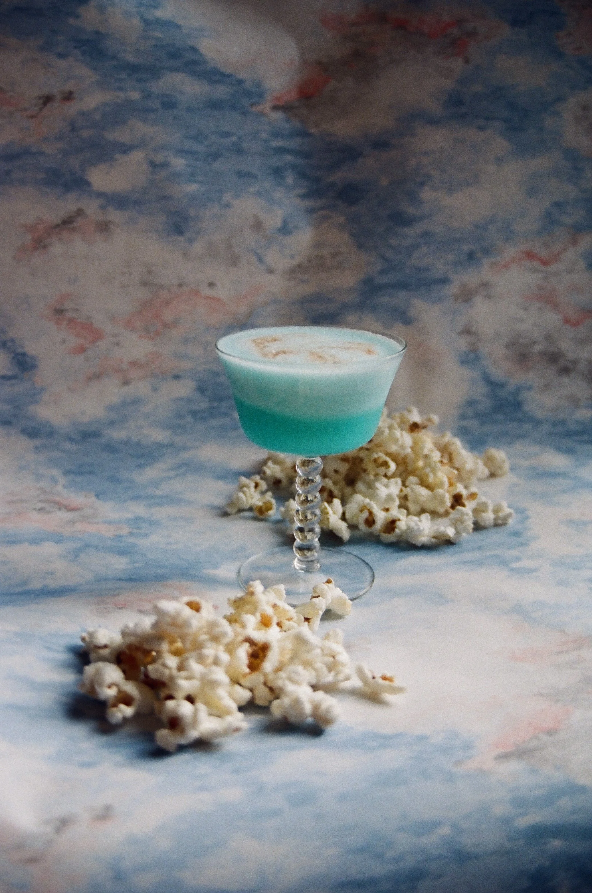 A teal and white layered cocktail in a fancy glass with a beaded stem, surrounded by scattered popcorn, against a cloudy sky backdrop.