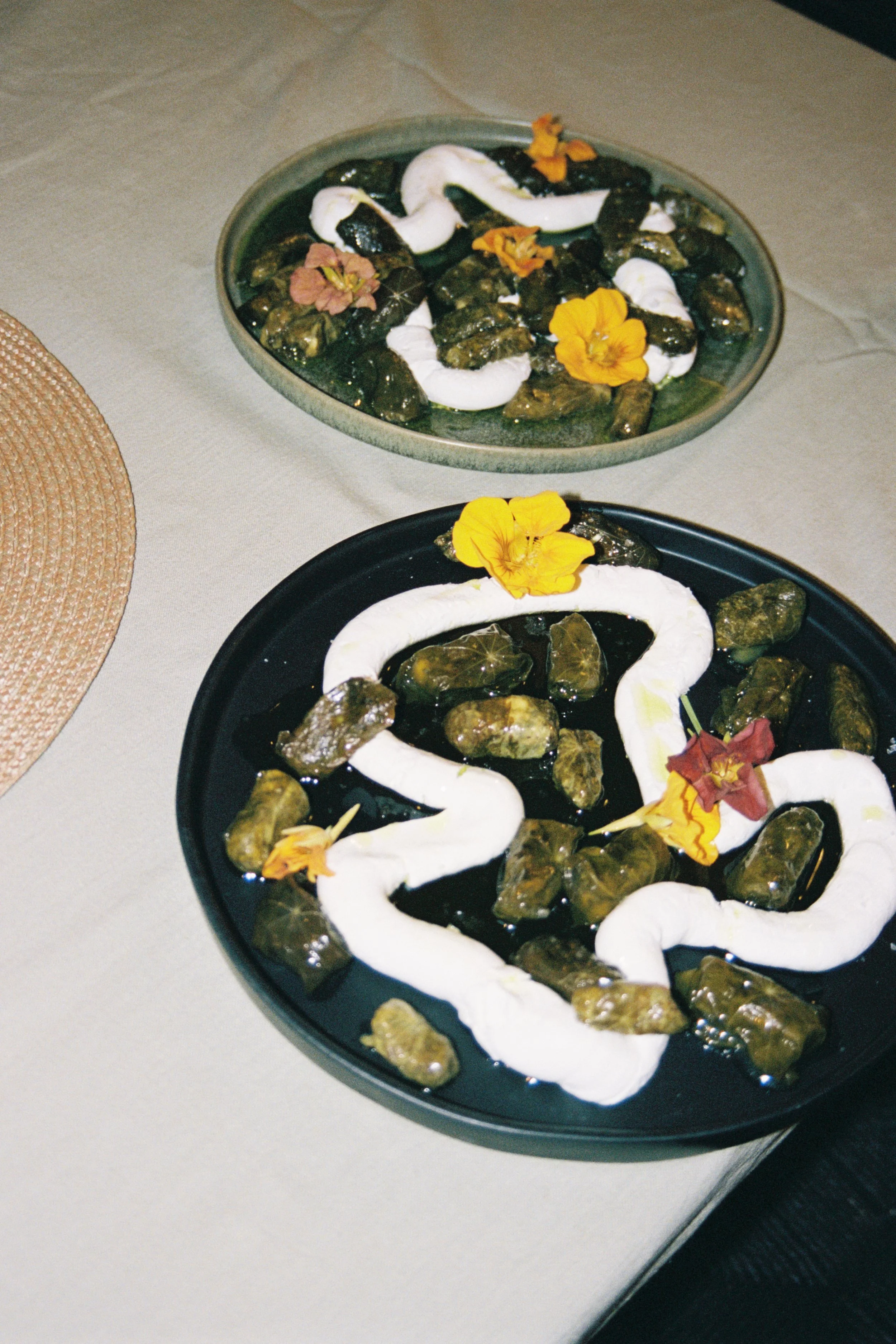 Two black plates filled with palm-sized sticky rice cakes topped with white swirls of cream and edible flowers, placed on a beige tablecloth.