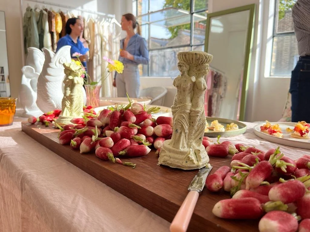 A table covered with a pink cloth holding red and white radish produce, decorative ceramic figurines, plates with appetizers, and glassware. Two women are engaged in conversation in the background near a window, with clothing hanging on a rack behind
