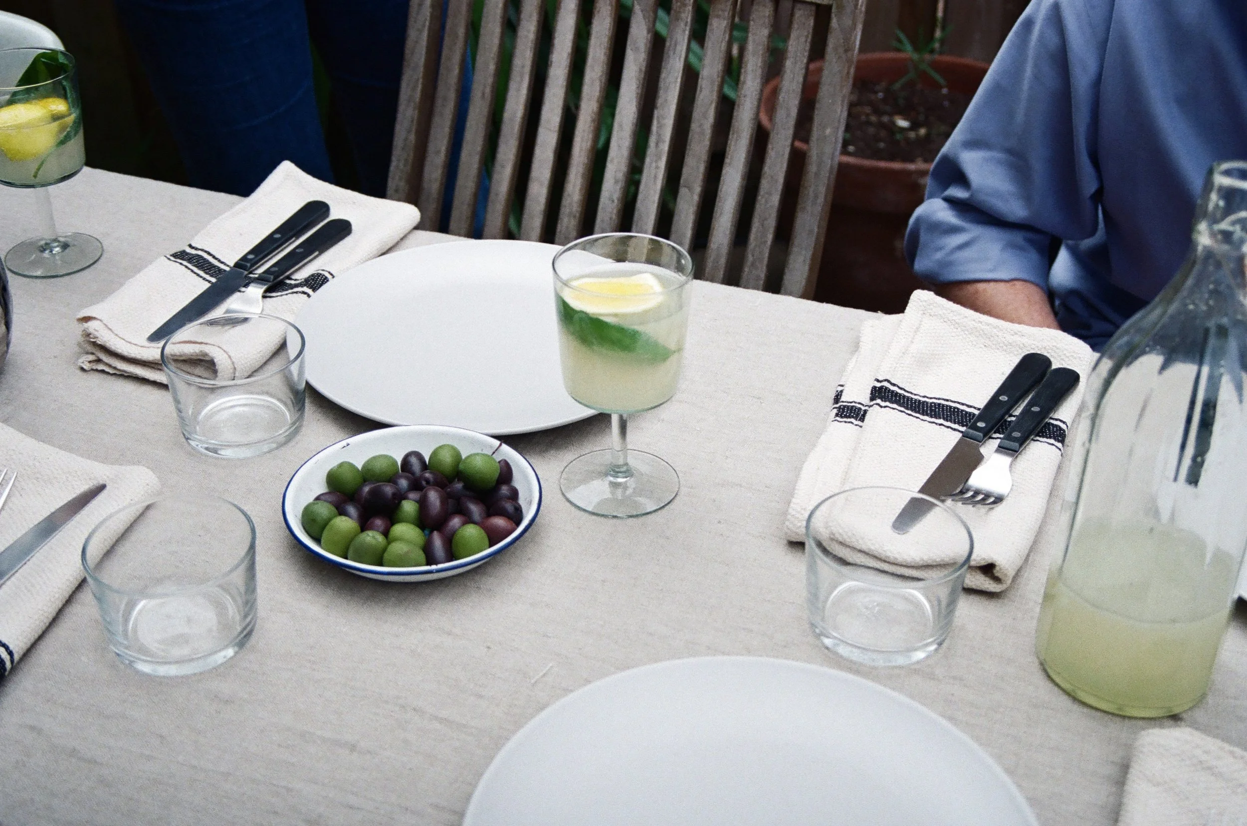 A table set outdoors with empty white plates, drinking glasses, knives and forks on napkins, a bowl of green and black olives, a glass of lemonade with lemon and mint, and a large water pitcher, with chairs and plants in the background.
