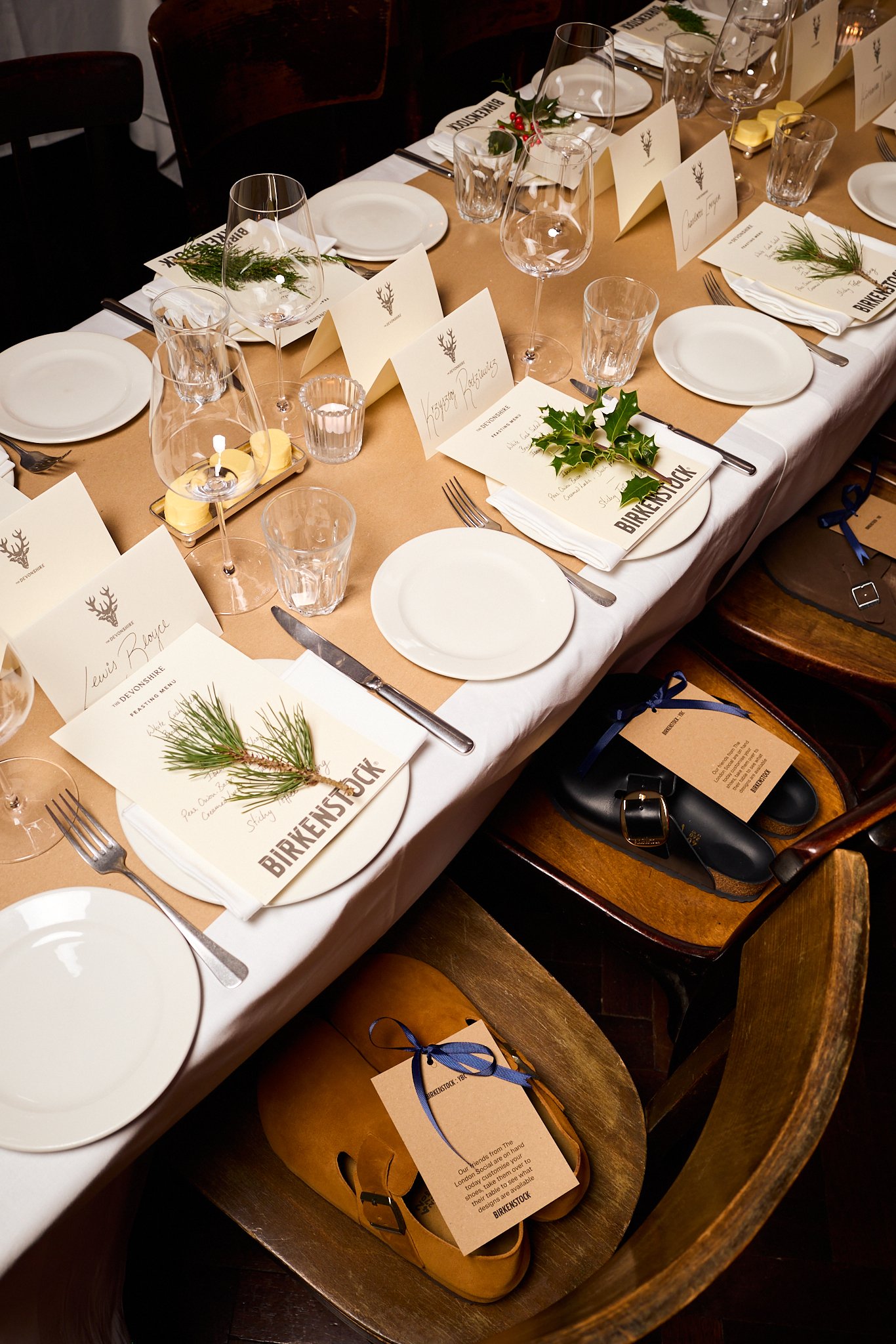A festive table setting with white plates, silverware, clear wine glasses, and rustic holiday decor including sprigs of pine and holly. Personalized name cards are placed at each setting, with some on top of folded napkins. There are candles and smal