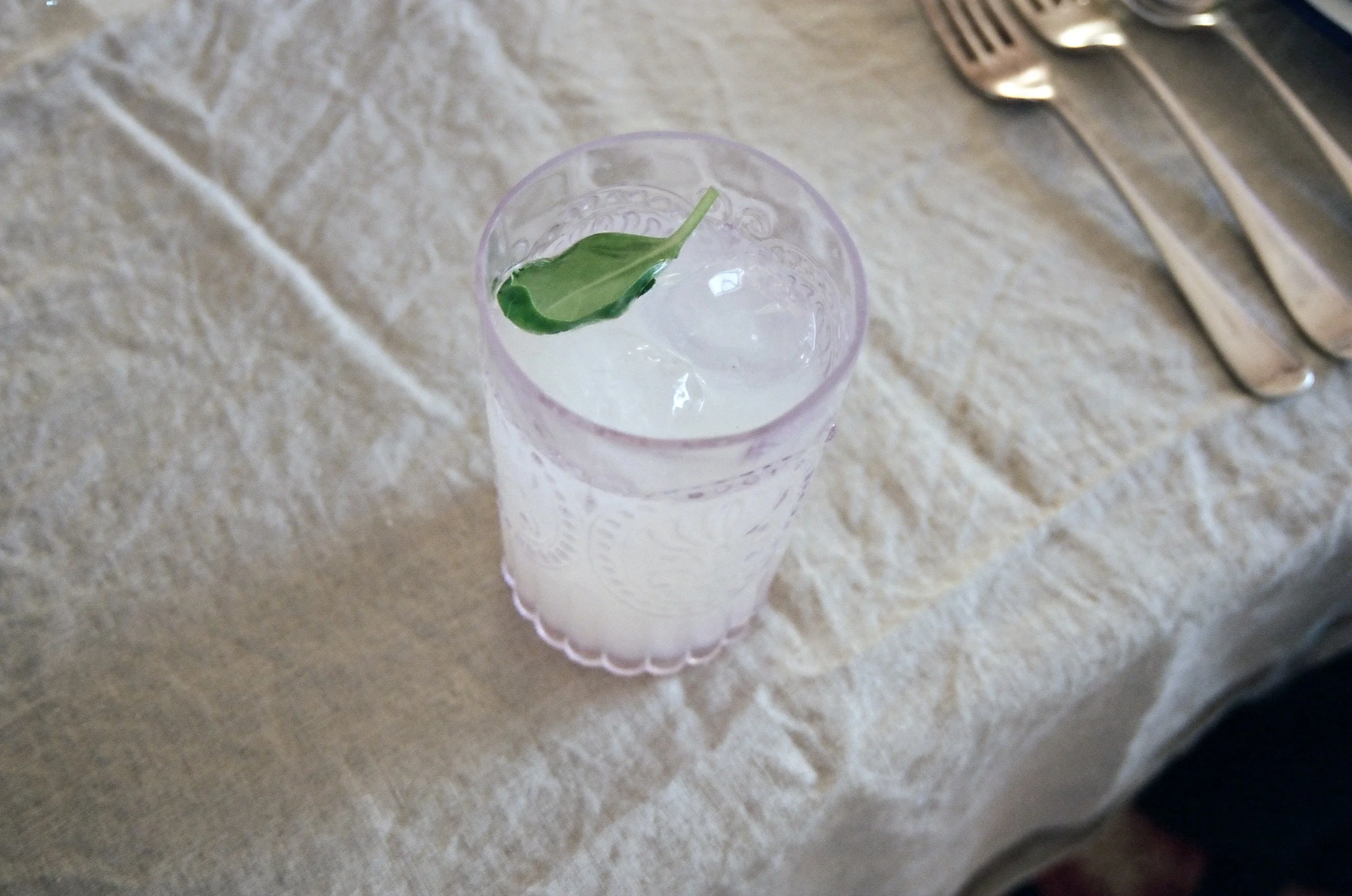 A tall glass with a light pink pattern contains a drink with ice cubes and a green leaf garnish, placed on a beige tablecloth near some silverware.