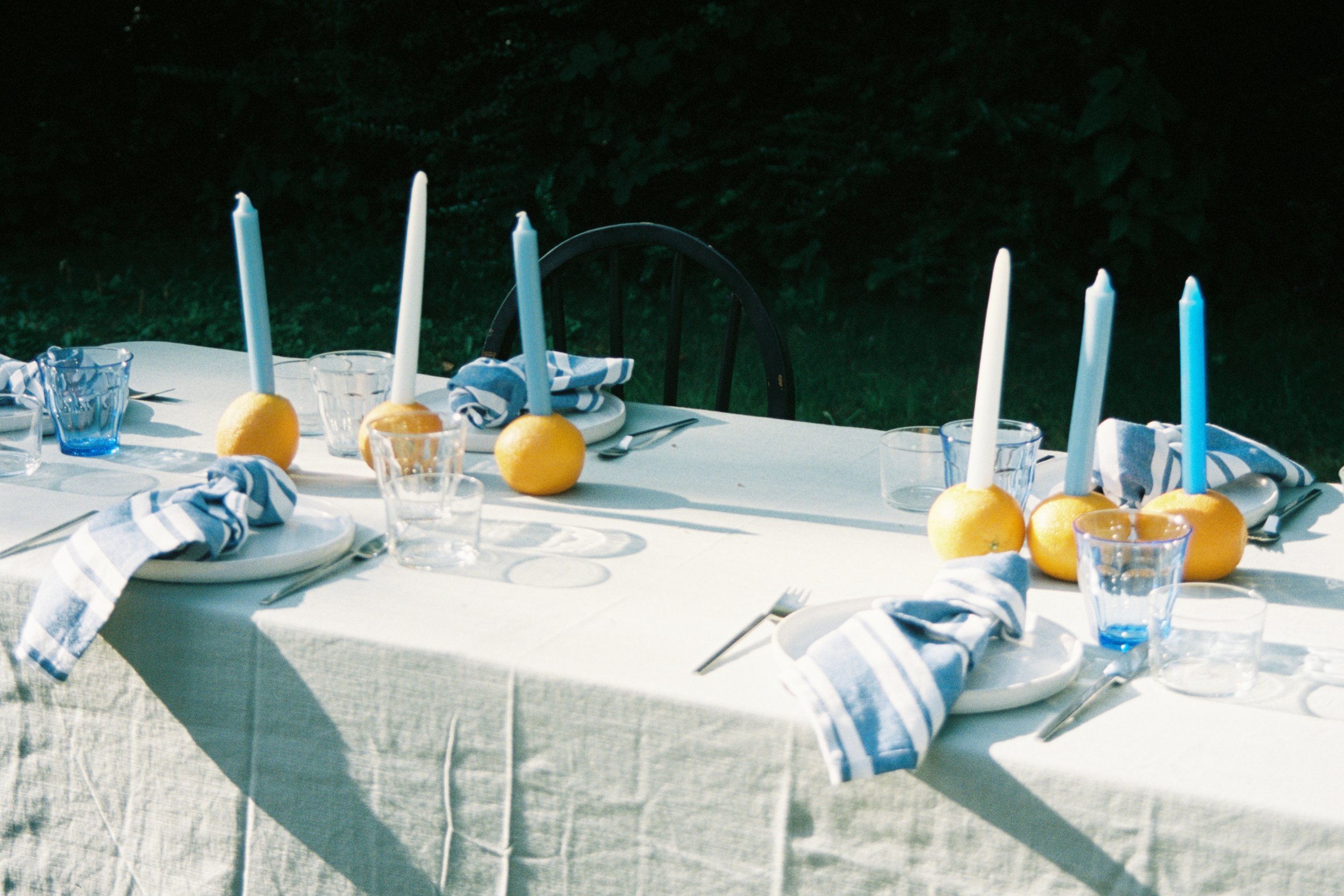 An outdoor dining table set for a meal with white candles in lemon holders, blue and white striped napkins, clear glassware, and white plates, with a garden in the background.