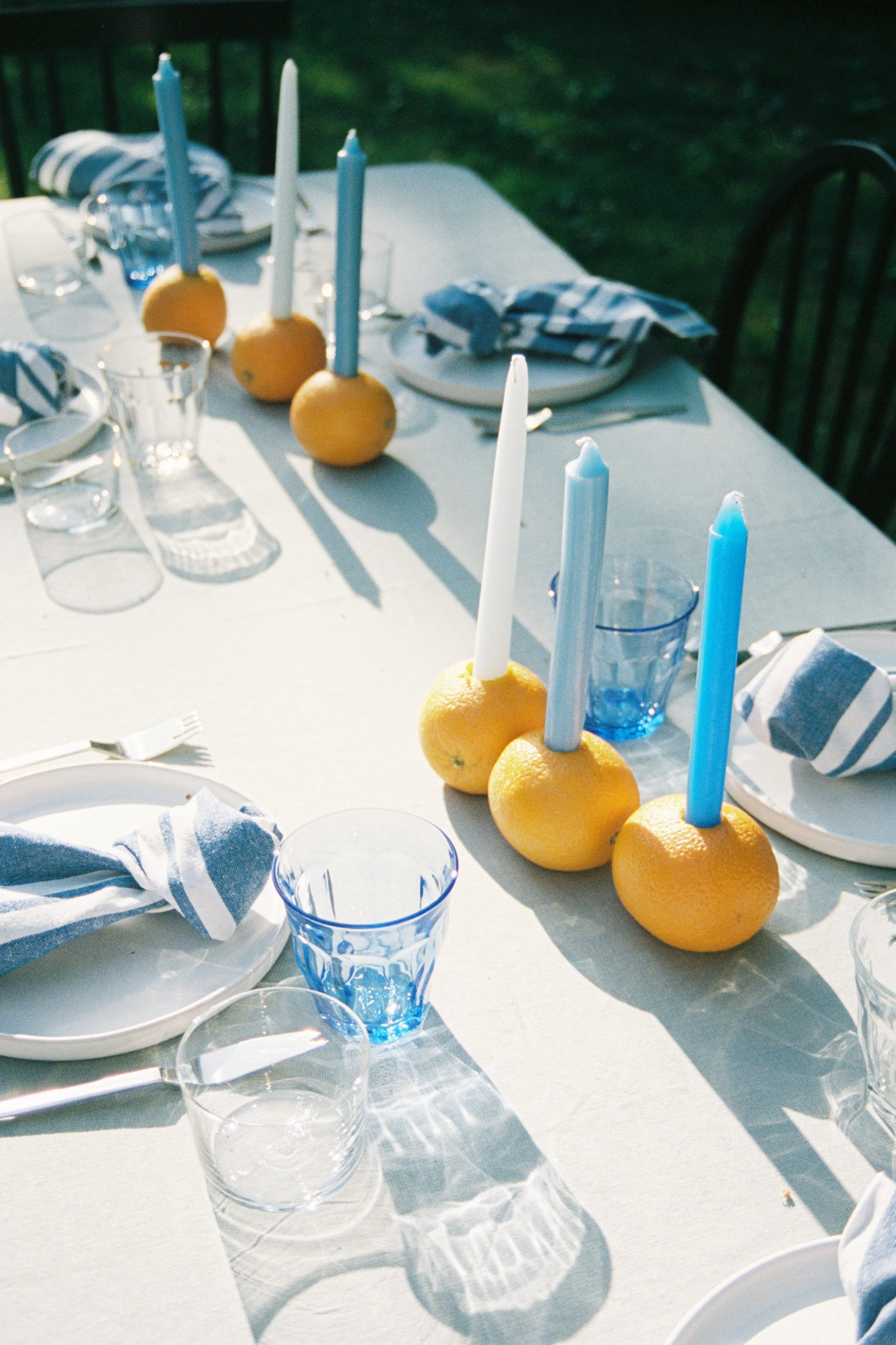 Table set for outdoor meal with blue and white striped napkins, clear glassware, white plates, and candles placed in citrus fruits, with a background of green grass.