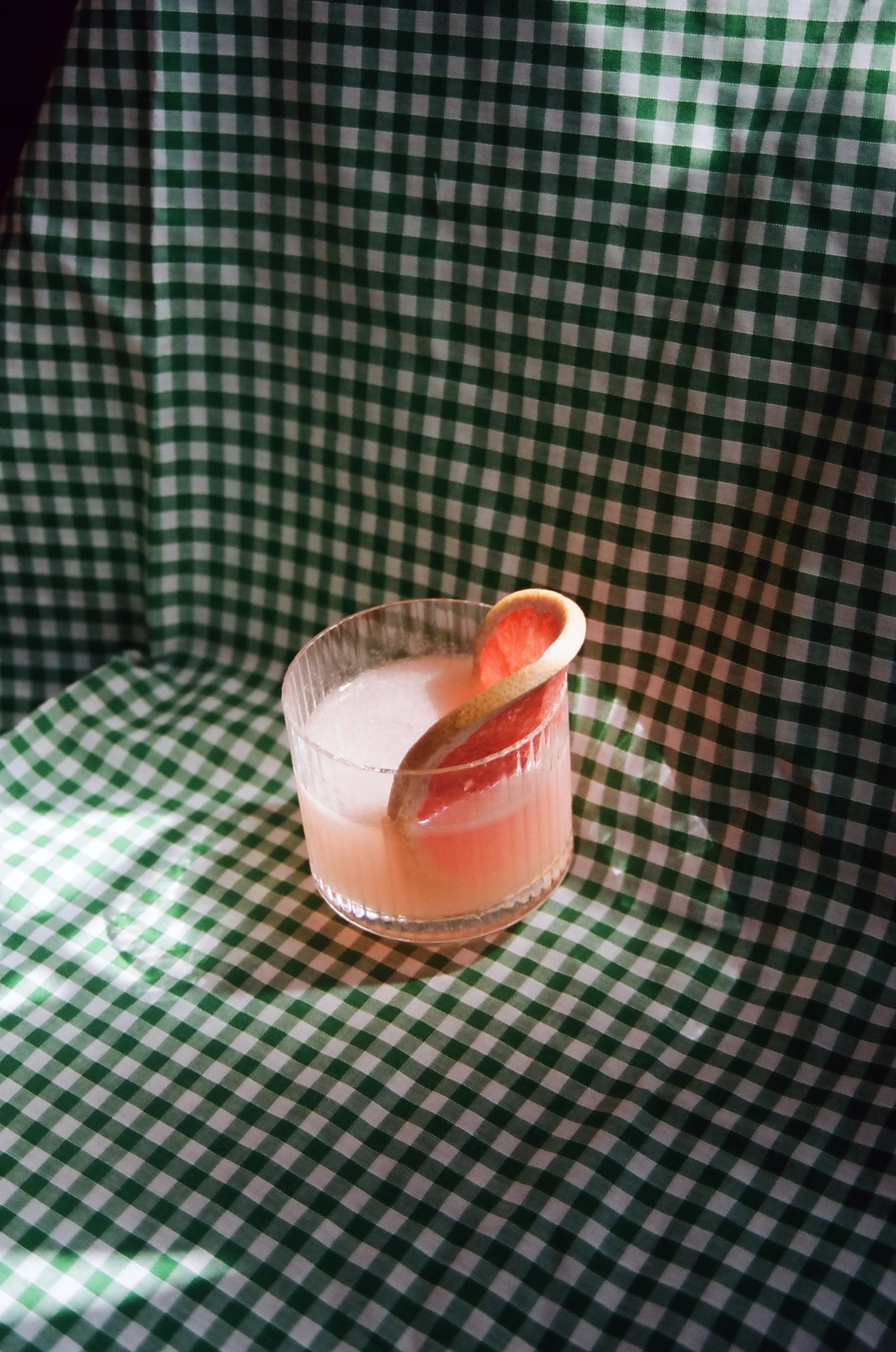 A glass of pink grapefruit juice with a slice of grapefruit as garnish on a green and white checkered tablecloth.