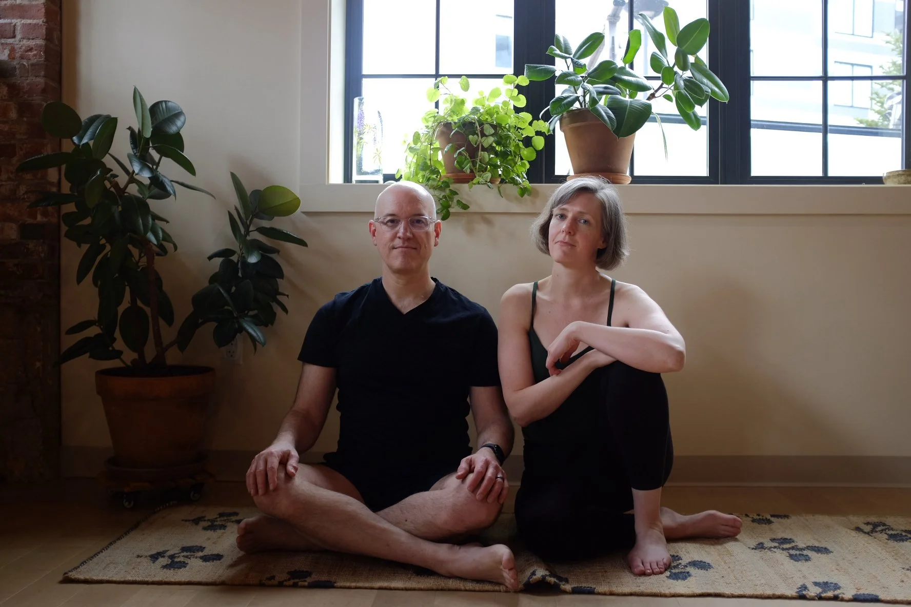 A man and a woman sitting cross-legged on a beige rug indoors, with potted plants behind them near a window.