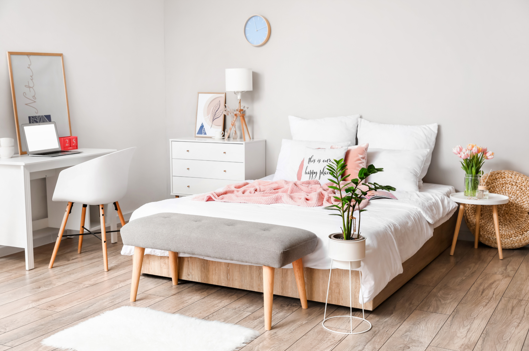 A bright, modern bedroom with a white bed, pink and white pillows, and a pink throw blanket. There is a small white side table with a glass vase of pink tulips, and a gray upholstered bench at the foot of the bed. A white dresser with artwork, a lamp, and decorative items is next to the bed, and a blue wall clock hangs above. To the left, a white desk with a white chair, a laptop, and decorative items is visible. The room has light wood flooring and minimal, cozy decor.