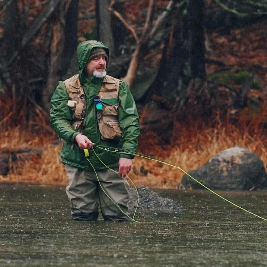 A man in rain gear standing waist-deep in a river, fishing with a line in an outdoor forest setting.