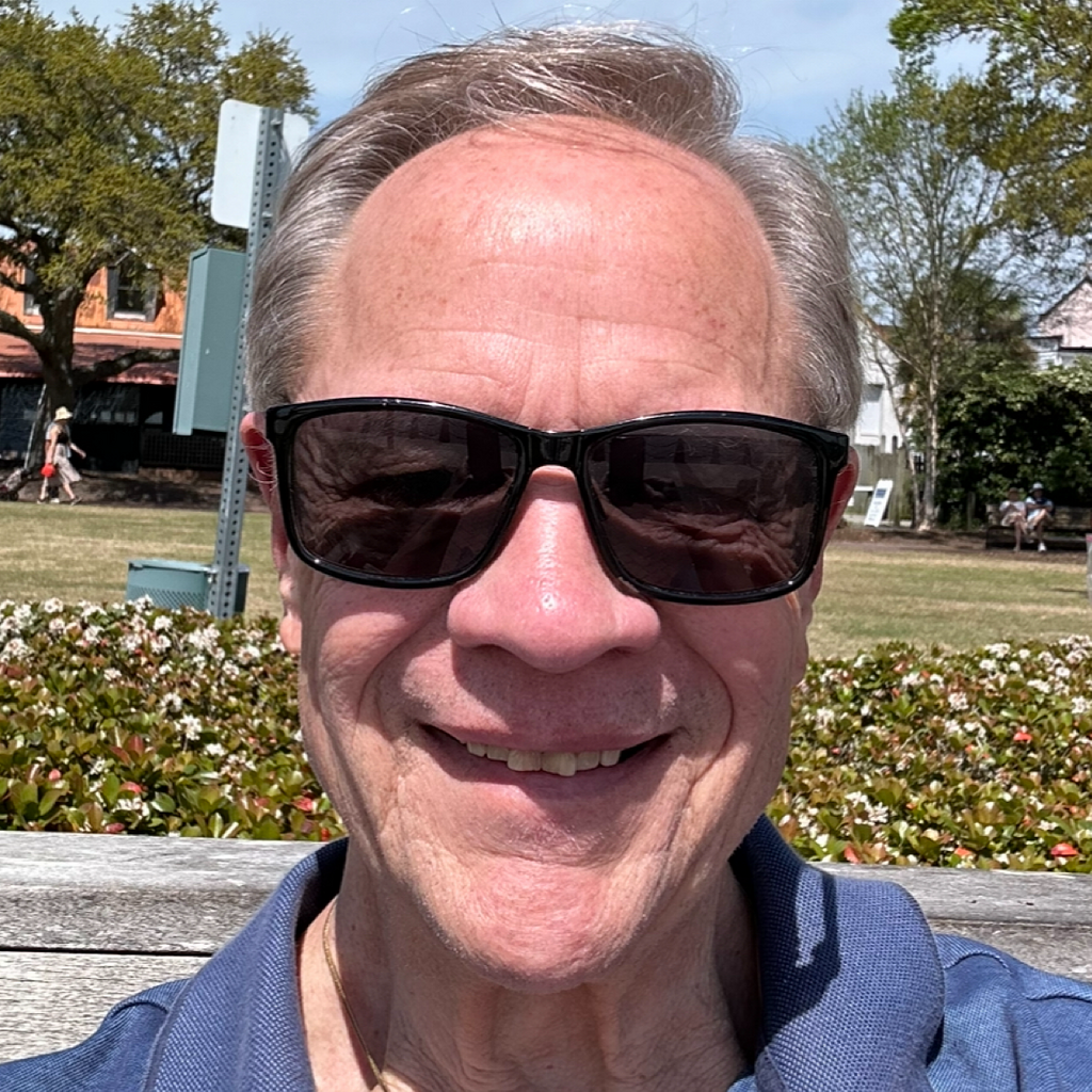 A smiling older man wearing sunglasses takes a selfie outdoors in a park with trees, bushes, and a bench.