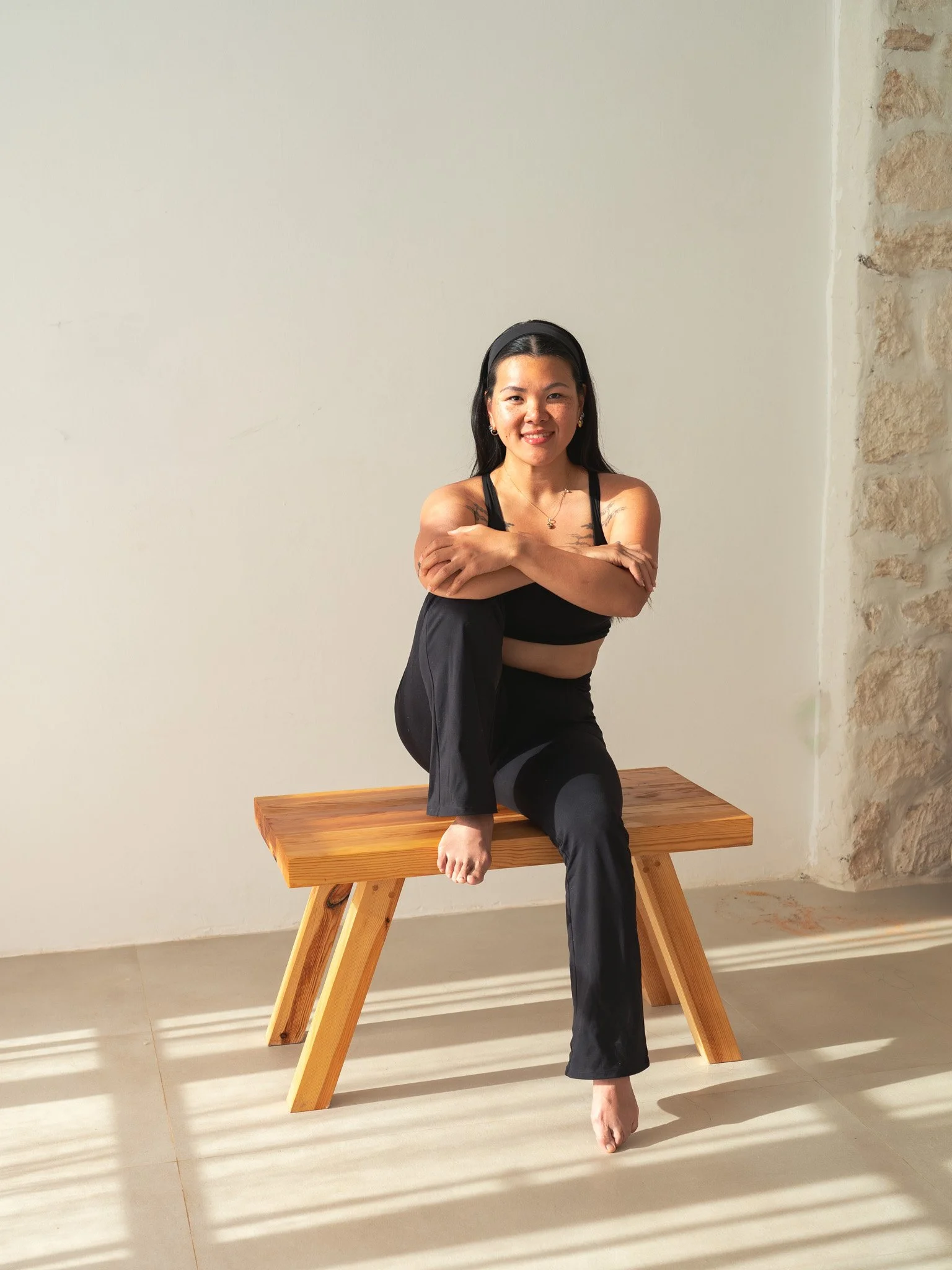 A woman sitting on a wooden bench crossing her arms, wearing a black sports bra and black pants, smiling at the camera in a minimally decorated room with beige tile floors and a plain white wall.