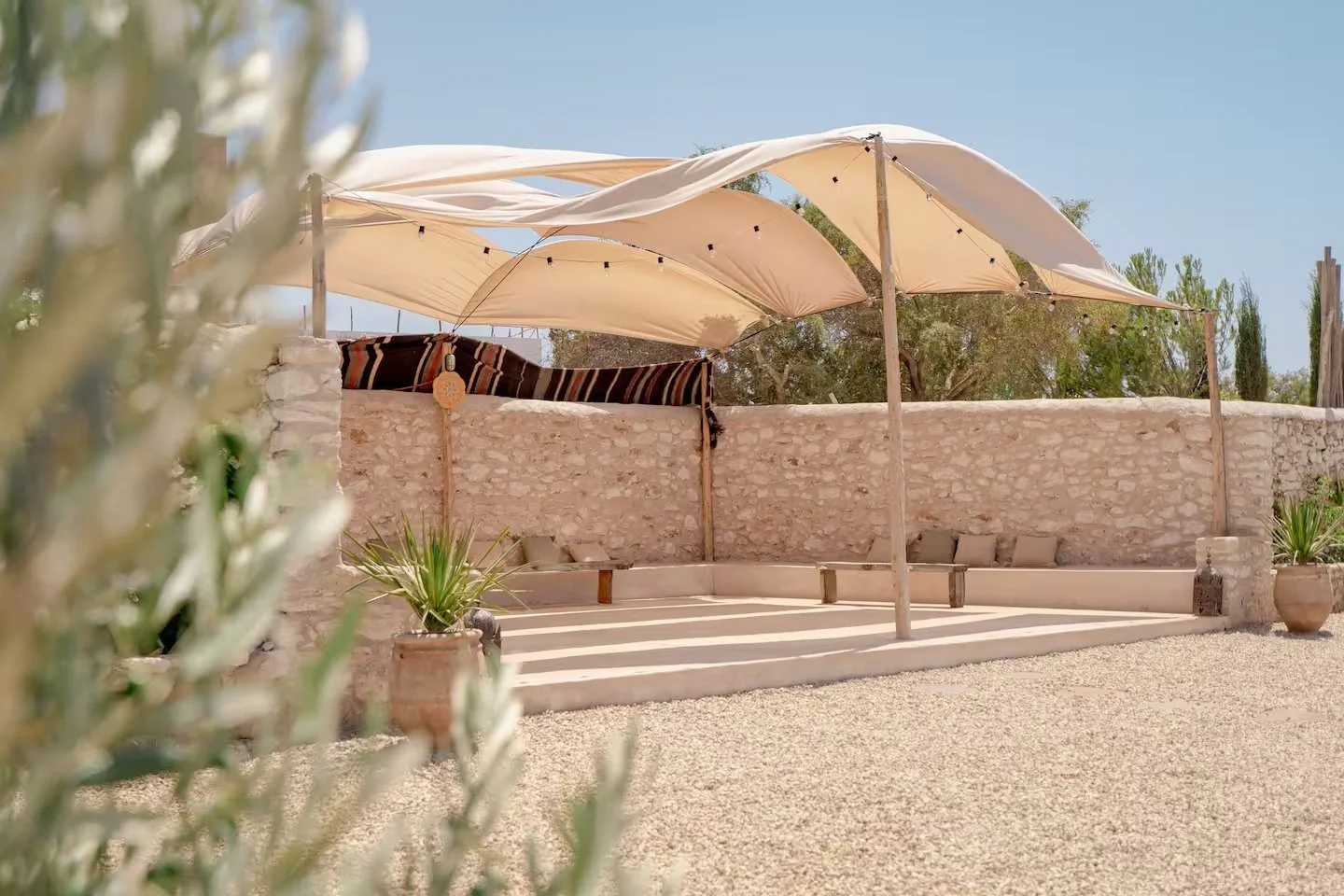 Outdoor seating area with a stone wall, covered by large white umbrellas, surrounded by potted plants and cushions, under a clear blue sky.