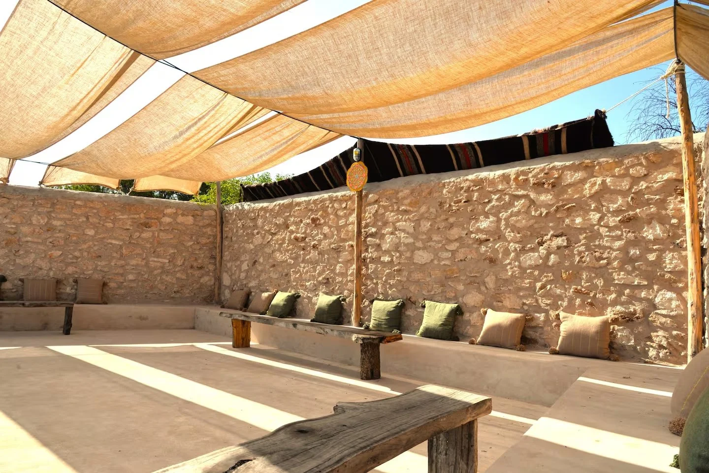 Outdoor seating area with stone wall, beige shade sails, wooden benches with cushions, and a decorative hanging lantern.