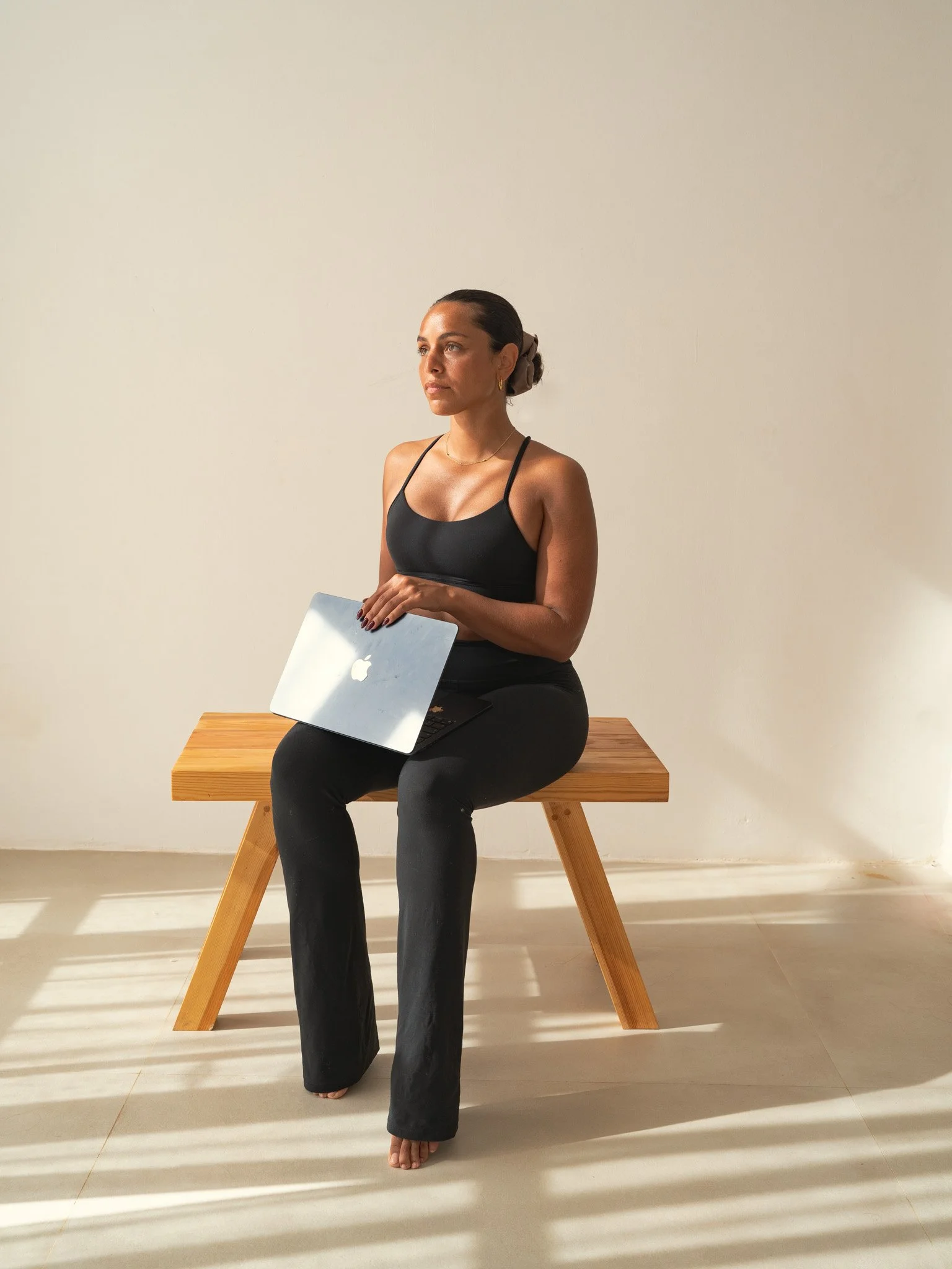 A woman sitting on a wooden bench with a laptop on her lap, against a plain light-colored wall.