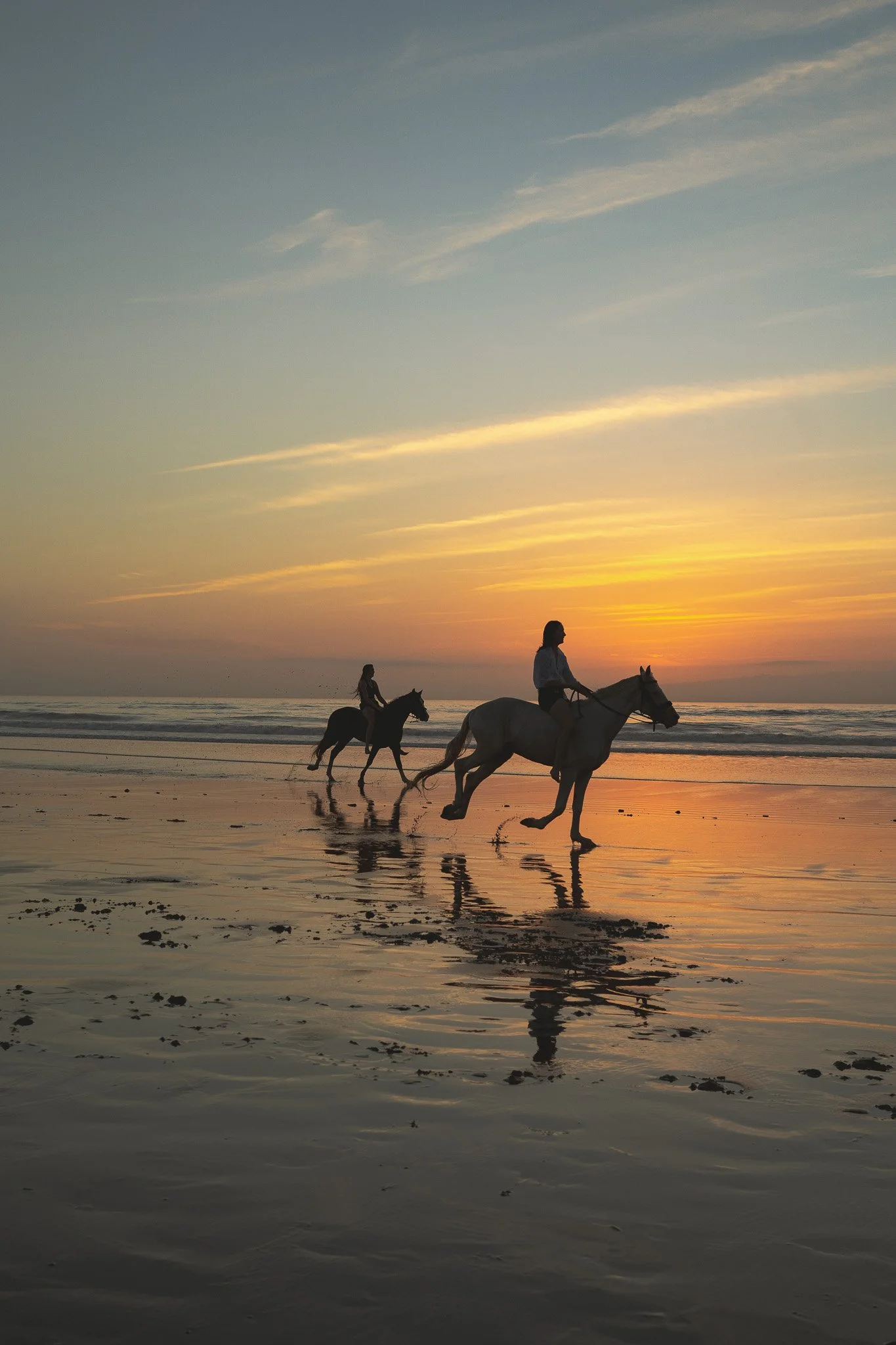 Two people riding horses along the beach at sunset, with the sky painted in orange and yellow hues and their reflections visible on the wet sand.
