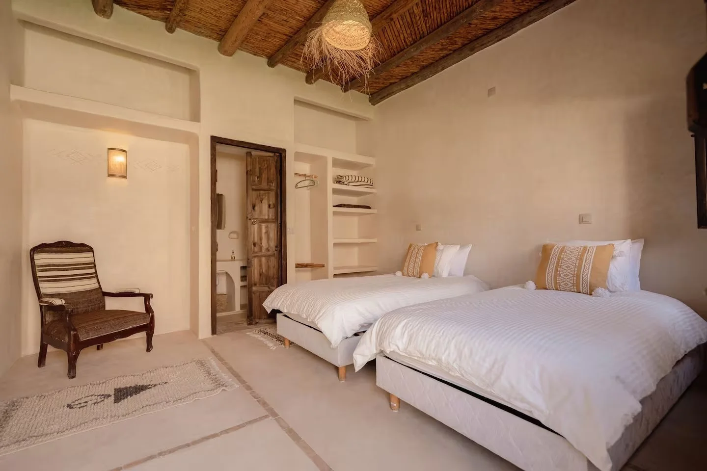 A bedroom with two single beds, a wooden armchair, built-in shelves, and a rustic wooden door. The room has a neutral color palette, textured ceiling, and decorative pillows.