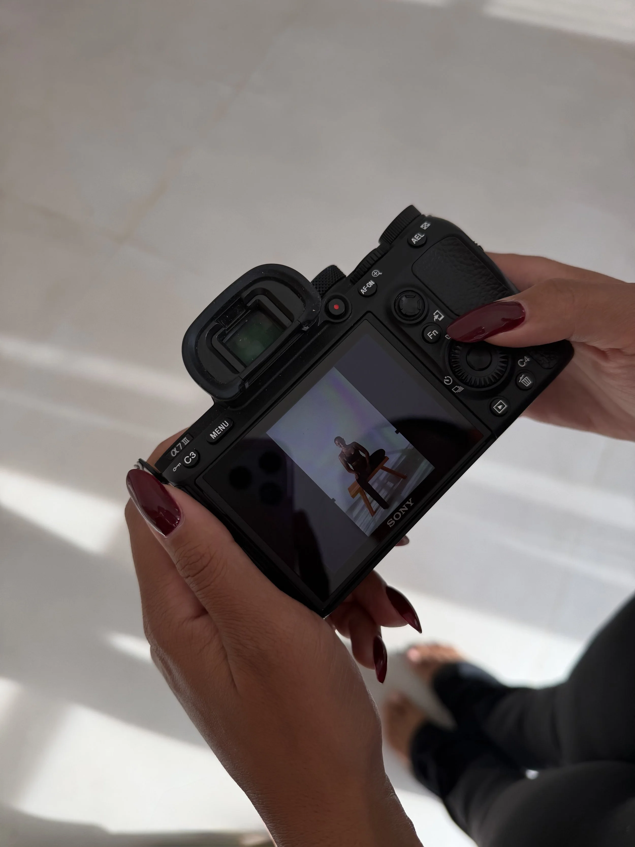 A person holding a Sony camera, capturing an image of a person sitting on a stool against a white background.
