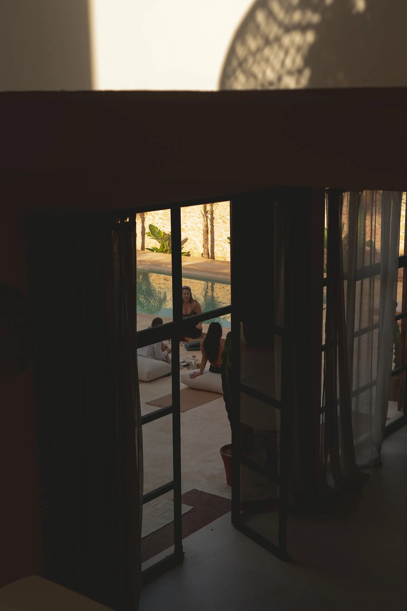 View through a window showing three women by a swimming pool, one of whom is sitting on the edge of the pool, in a relaxed setting with natural light.