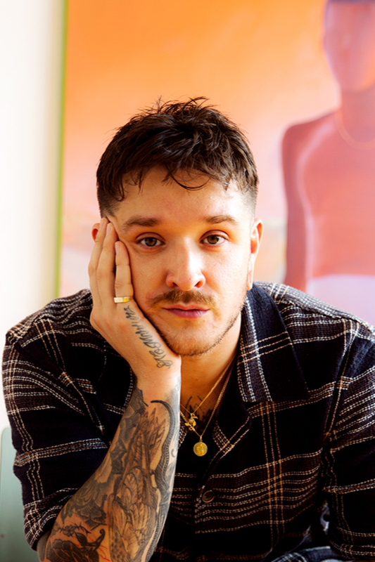 A young man with tattoos on his arm resting his head on his hand, wearing a black plaid shirt and gold jewelry, sitting indoors with colorful art in the background.