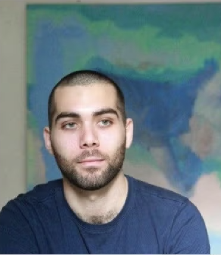 A man with short dark hair and a beard, wearing a navy blue shirt, sitting in front of a colorful map background.