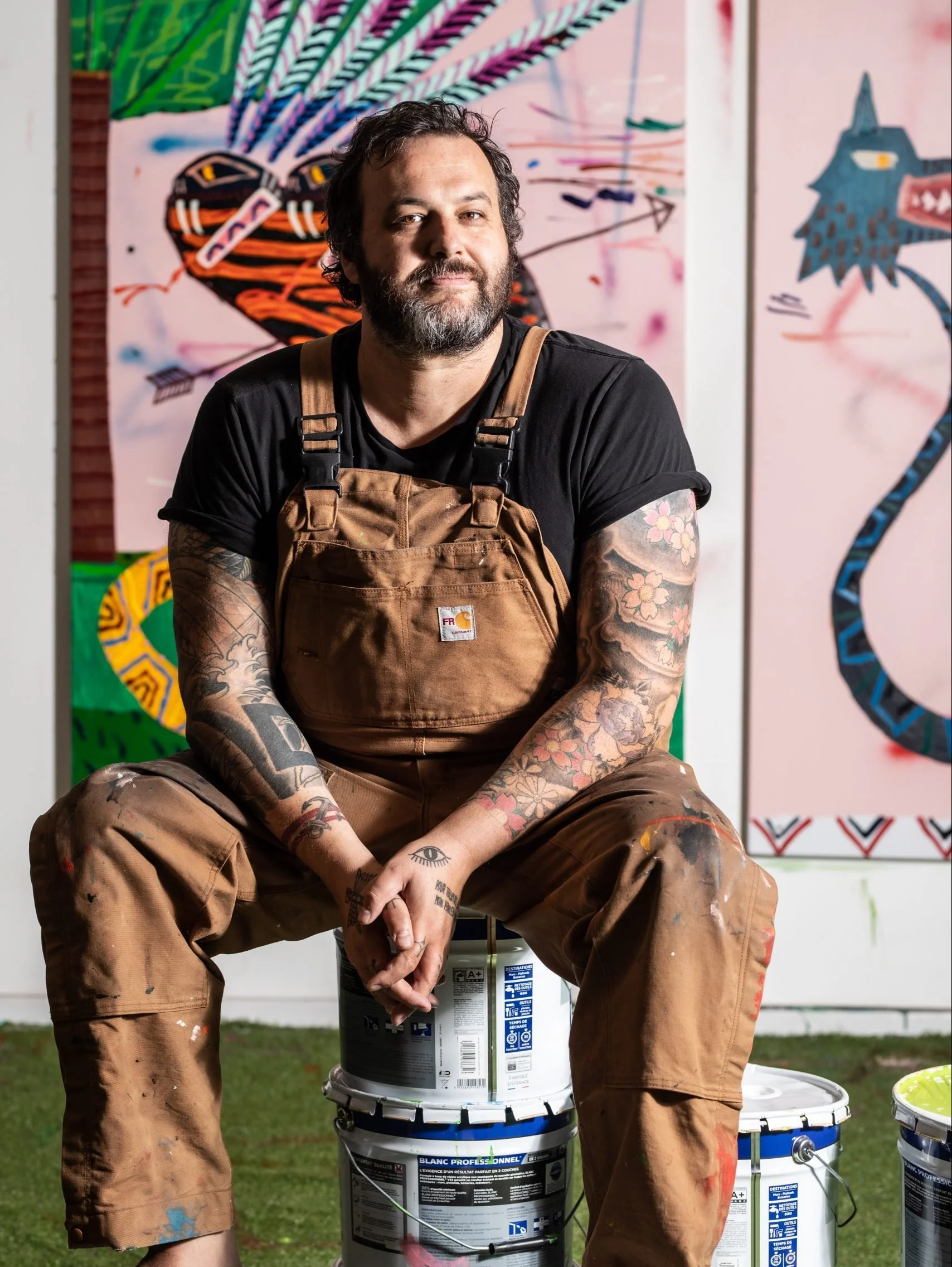 A man with tattoos, beard, and curly hair sits on paint buckets in front of colorful abstract artwork.