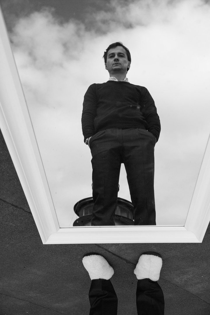 A man taking a photo of his reflection in a mirror placed on the ground, showing his feet wearing white fuzzy slippers and the man’s reflection looking down at the mirror