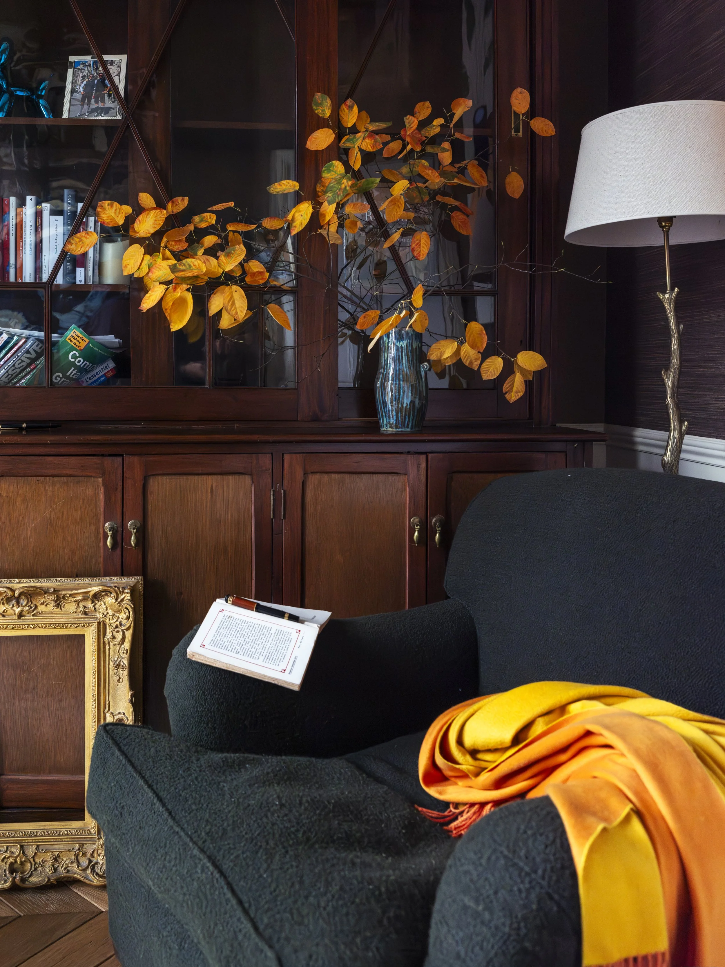 Living room with dark wood bookshelf, a vase with orange and yellow autumn leaves, a black armchair with an open book and a multicolored scarf, and a white lampshade.
