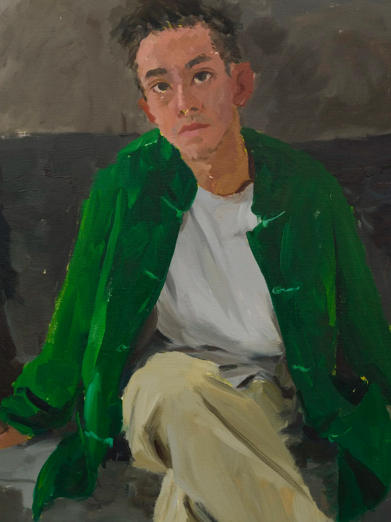A portrait of a young man with short dark hair, wearing a green jacket over a white shirt, sitting against a dark background.