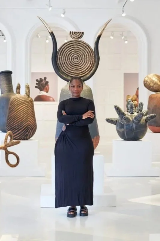 A woman in a black long-sleeve dress standing with crossed arms in an art gallery surrounded by large abstract sculptures.