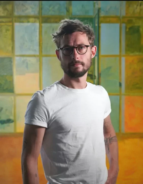 A man with glasses, tousled hair, and a beard wearing a white T-shirt standing in front of a colorful stained glass window.