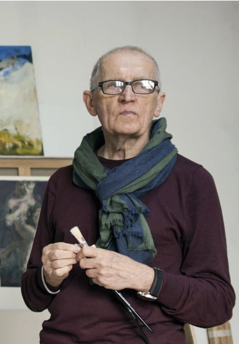 An elderly man with glasses, short gray hair, and fair skin stands indoors, holding a small paintbrush. He wears a maroon sweater and a multicolored scarf. Behind him are paintings on display.