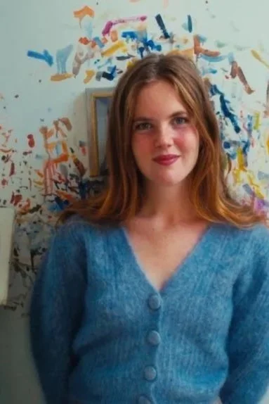 A young woman with red hair and wearing a blue sweater standing in front of a colorful, abstract painted wall.